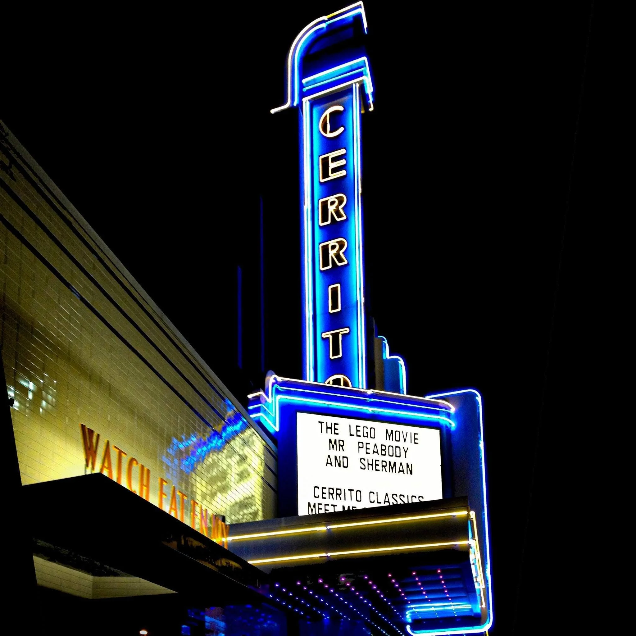 Exterior of a vintage theater with a marquee and a digital sign displaying showtimes and messages, surrounded by trees and storefronts, the exterior of El Cerrito.