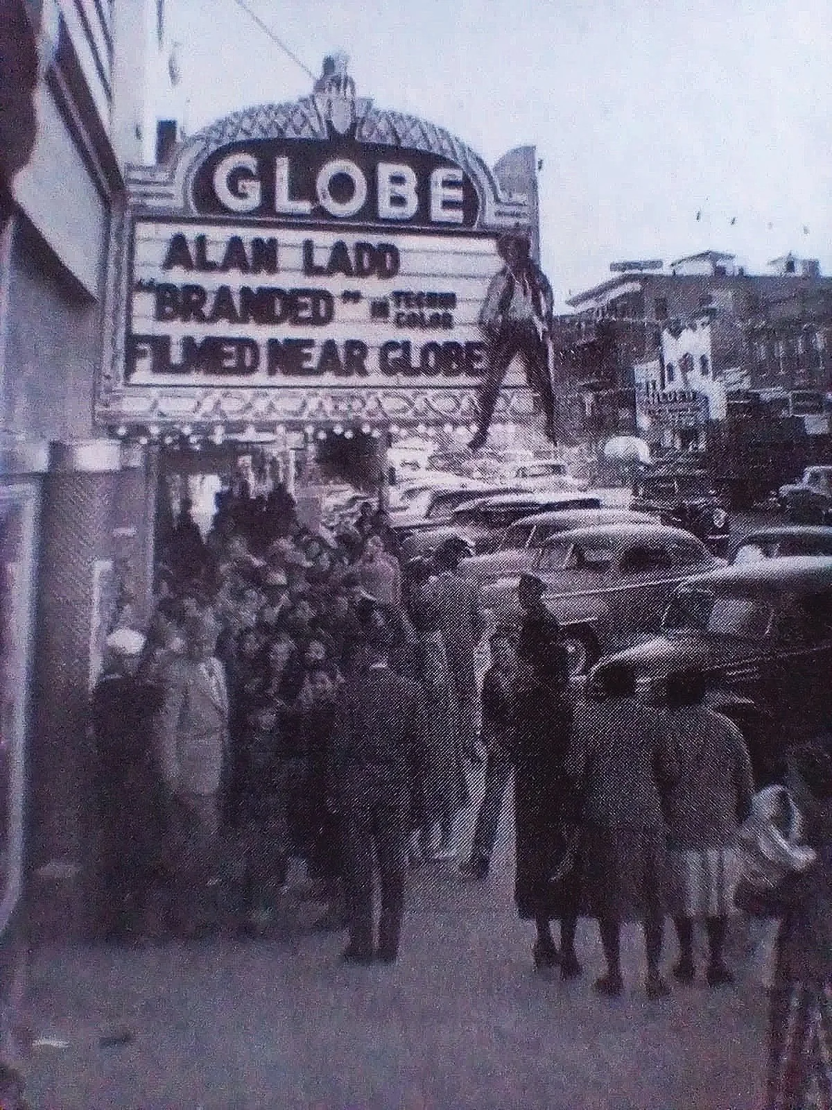 Black and white photo of a crowd outside a theater with a marquee sign that reads "GLOBE" and "ALAN LADD "BRANDED" IN TECHNICOLOR" and "FILMED NEAR GLOBE"