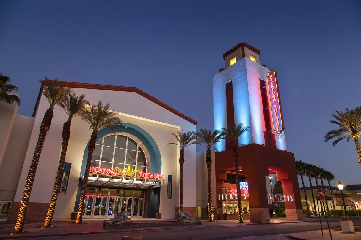 Night view of the Mary Pickford theater building, lit in blue and red neon lights, palm trees lining the street.