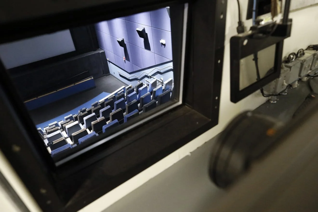 View through the porthole of the control room of Gateway Film Center, showing rows of seats, the screen, and wall-mounted speakers of the screening room below.
