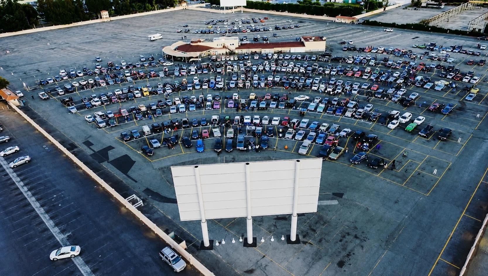 View of the Paramount Drive-in filled with many cars and a few people walking around, taken from a high angle. There is the screen in the foreground and the projection structure with a curved roof in the background.