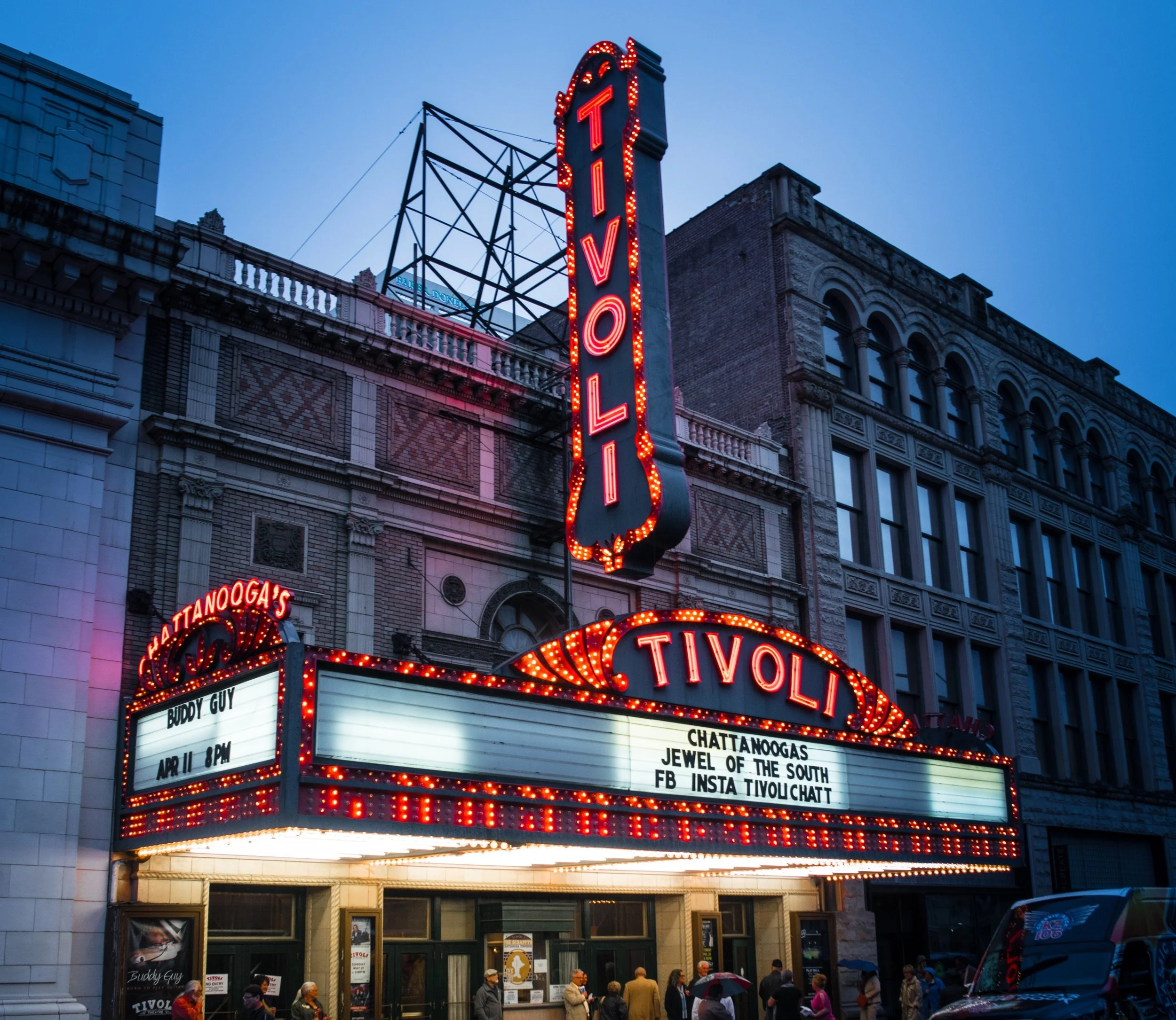 Nighttime view of the Tivoli Theatre marquee with bright red neon lights, surrounded by historic brick and stone buildings, and people walking on the sidewalk below.