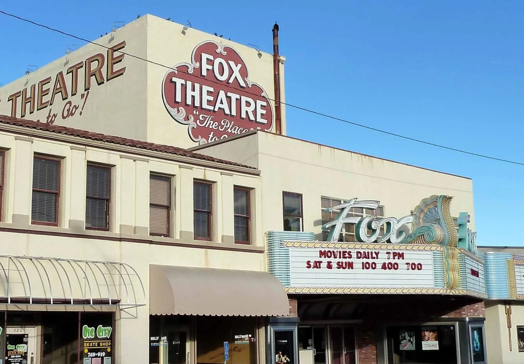 The Fox Theatre marquee sign displaying showtimes and movie schedule information, with part of the building's facade, windows, and a decorative sign visible against a clear blue sky.