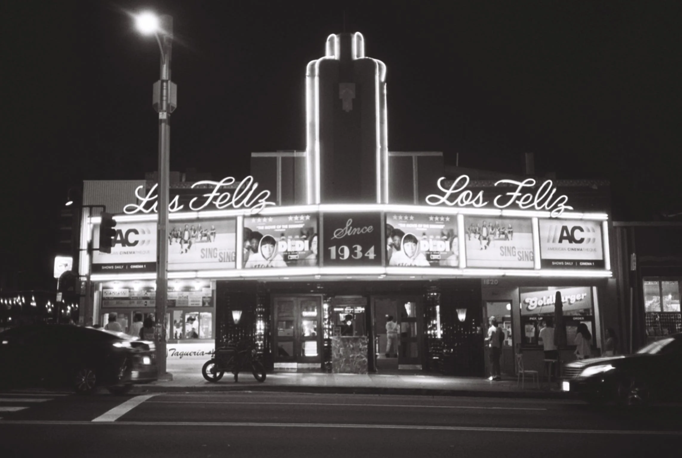 Nighttime scene of Los Felix 3 theater, advertising movies, with cars and pedestrians in front.