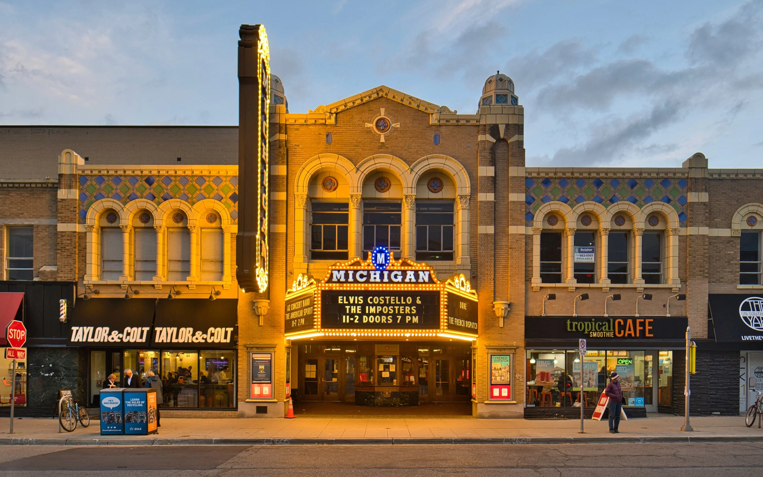 The Michigan theater building with a marquee advertising a concert by Elvis Costello & the Imposters. The building has a decorative brick facade with arched windows and illuminated signage. There are people walking and sitting outside on the sidewalk.