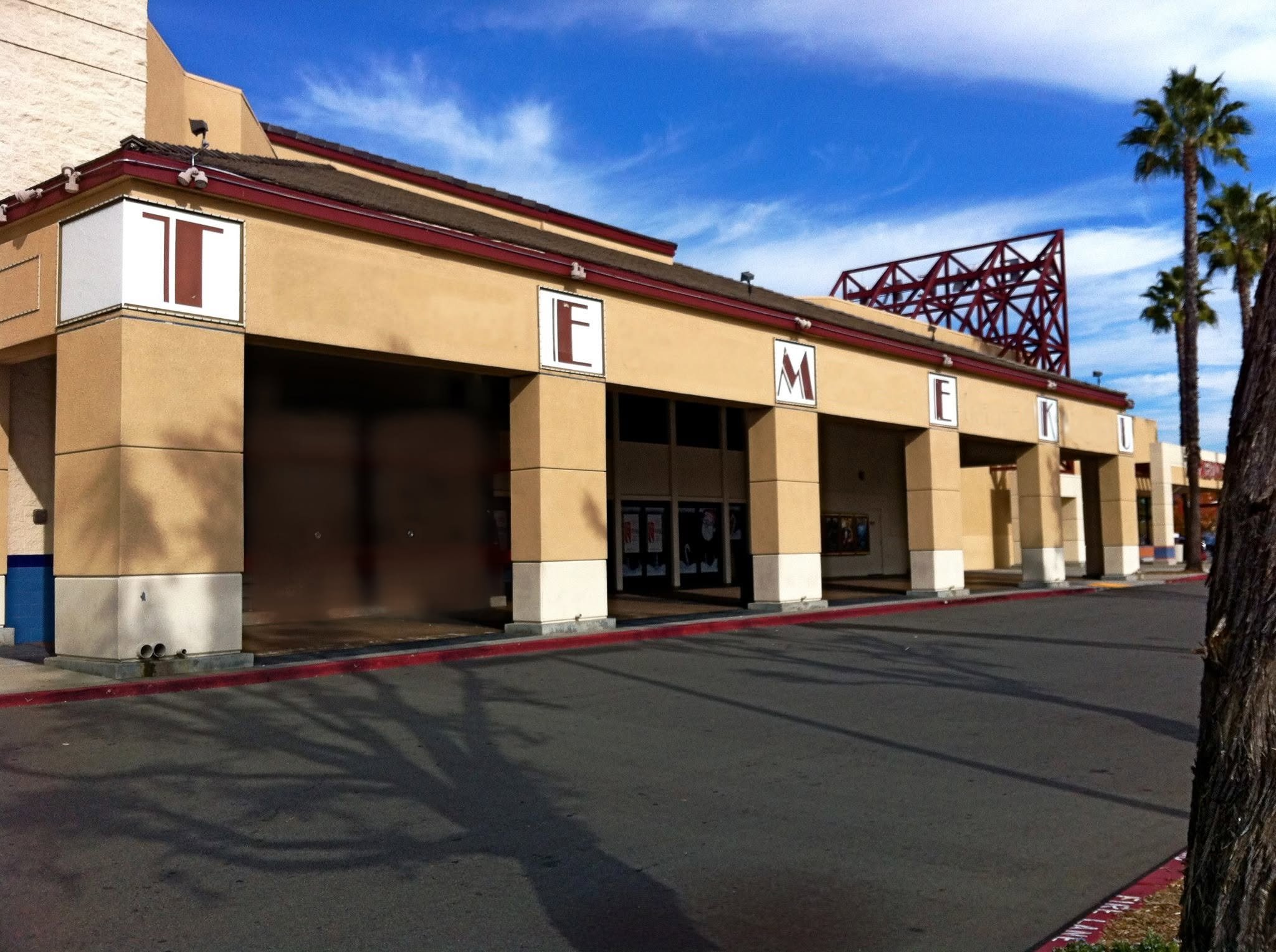 Exterior of a beige shopping mall with the word 'Temeku' in large individual letters on the facade, against a background of a partly cloudy blue sky and palm trees.