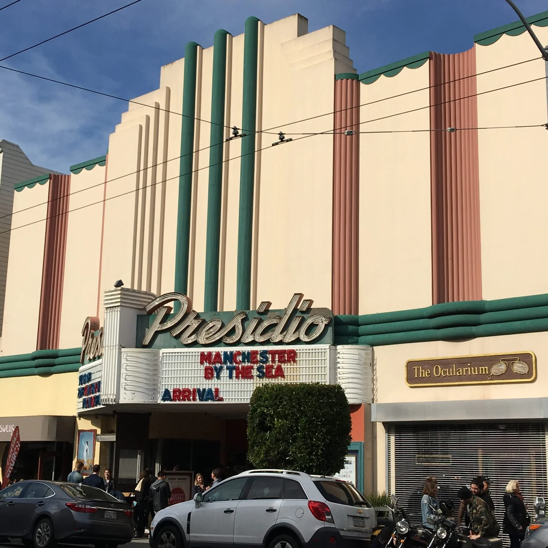 The facade of the Presidio, an independent movie theater with art deco architectural details including vertical green and brown columns and decorative scalloped edges under green awnings. People are outside and cars parked in front.