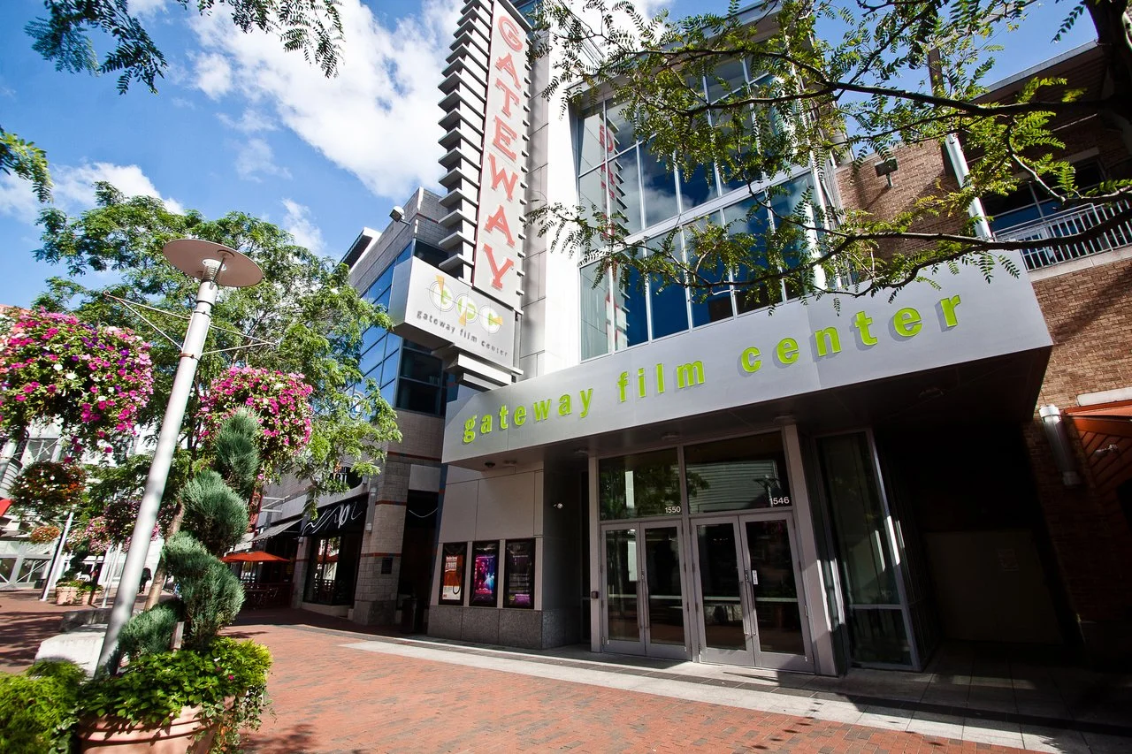 Exterior view of Gateway Film Center, a modern building with large glass windows, surrounded by trees and flowers, under a partly cloudy sky.