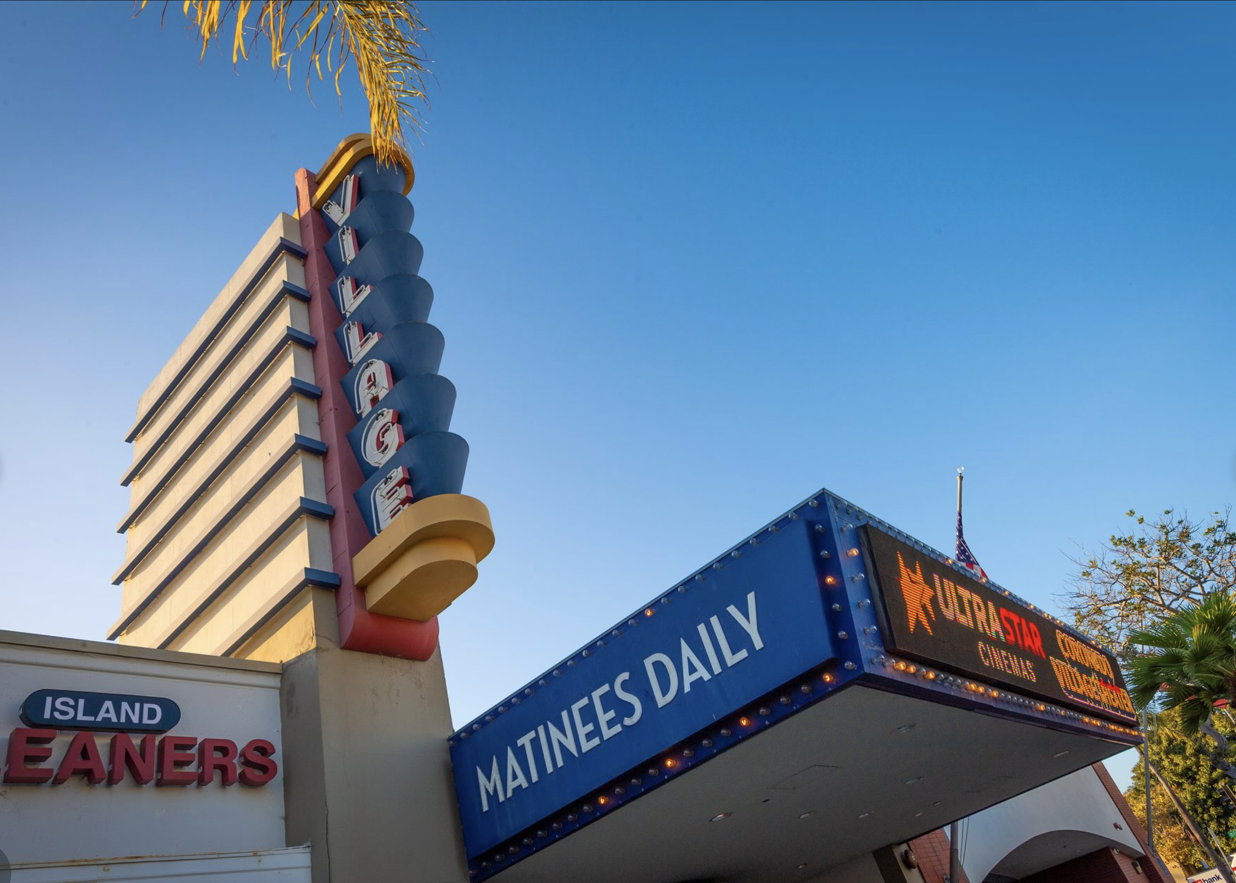 The colorful vertical of Village Theater in Coronado with stacked, rounded letters attached to the corner of the building, with part of an American flag visible behind trees and a clear blue sky.