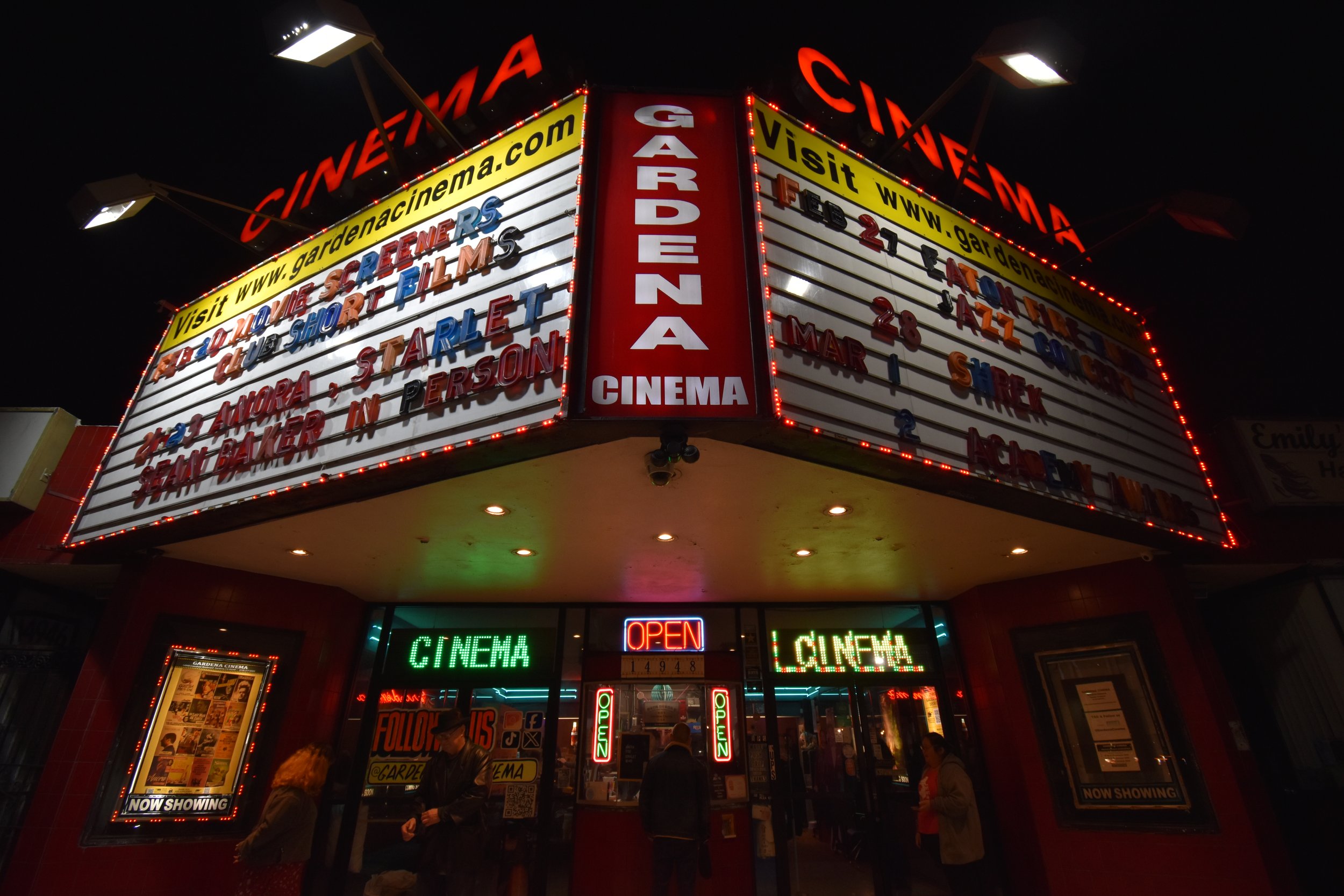 Night view of a vintage cinema theater entrance, the Gardena Cinema, with illuminated marquee signs, including the website www.gardencinemas.com and upcoming movie showings.