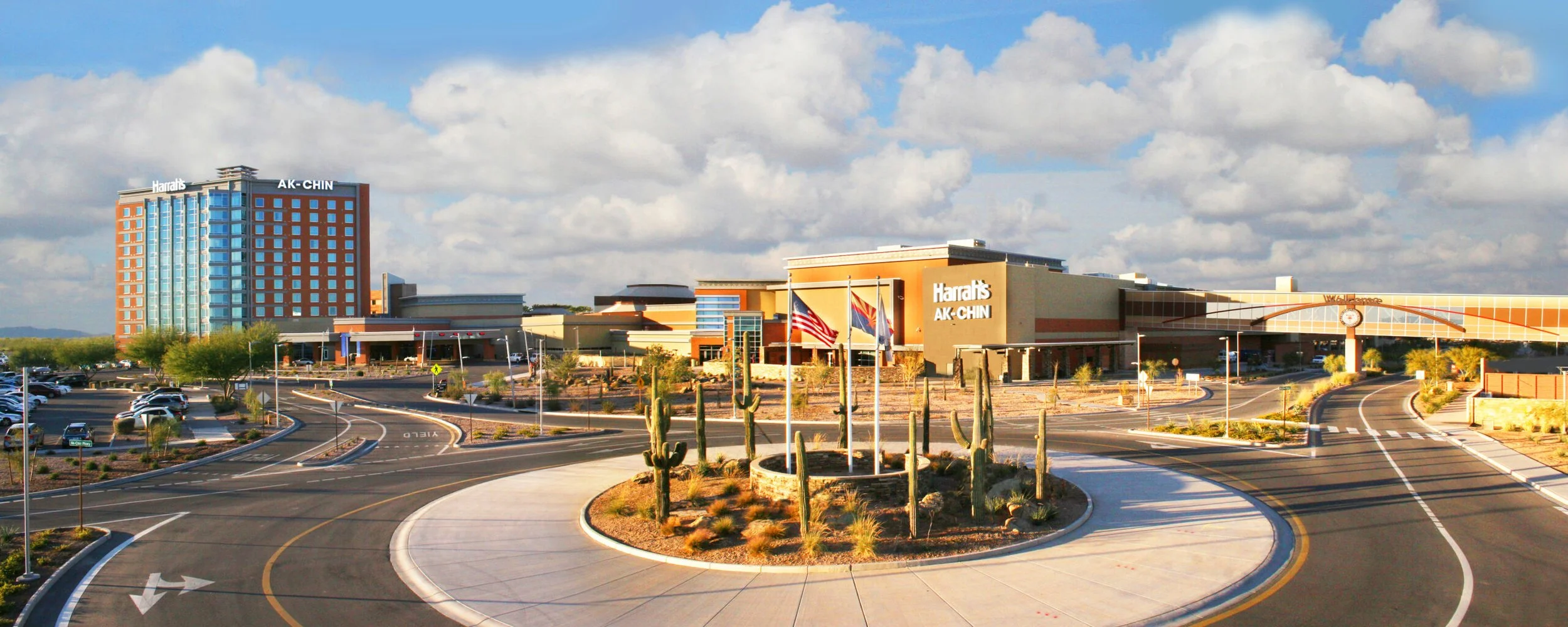 Parking lot and road leading to Harrah's AK-CHIN Casino resort with cactus plants in the foreground and a tall hotel building to the left, under a partly cloudy sky.