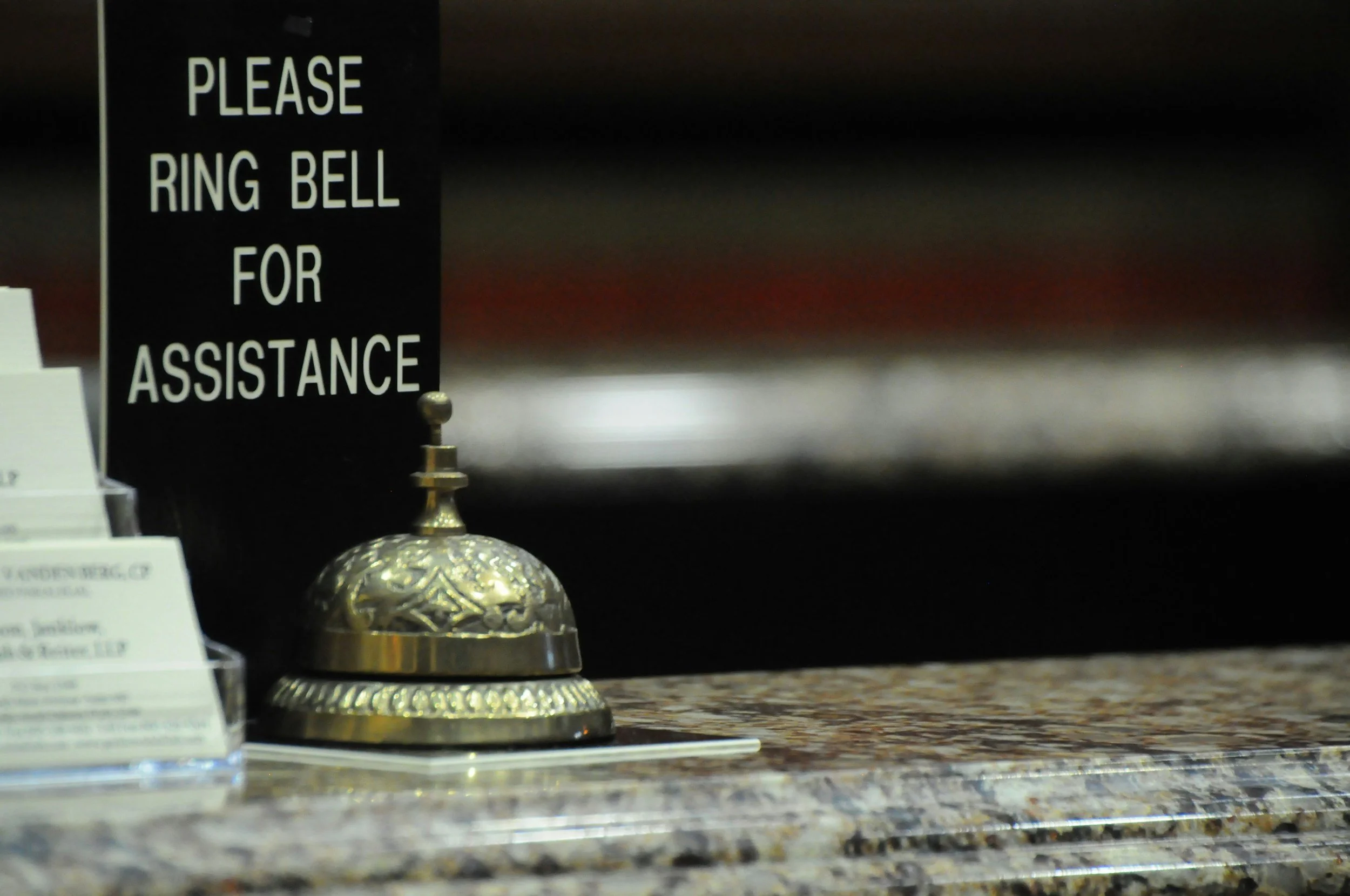 A black sign with white text that says, 'Please Ring Bell For Assistance,' placed on a decorative stand on a granite countertop.