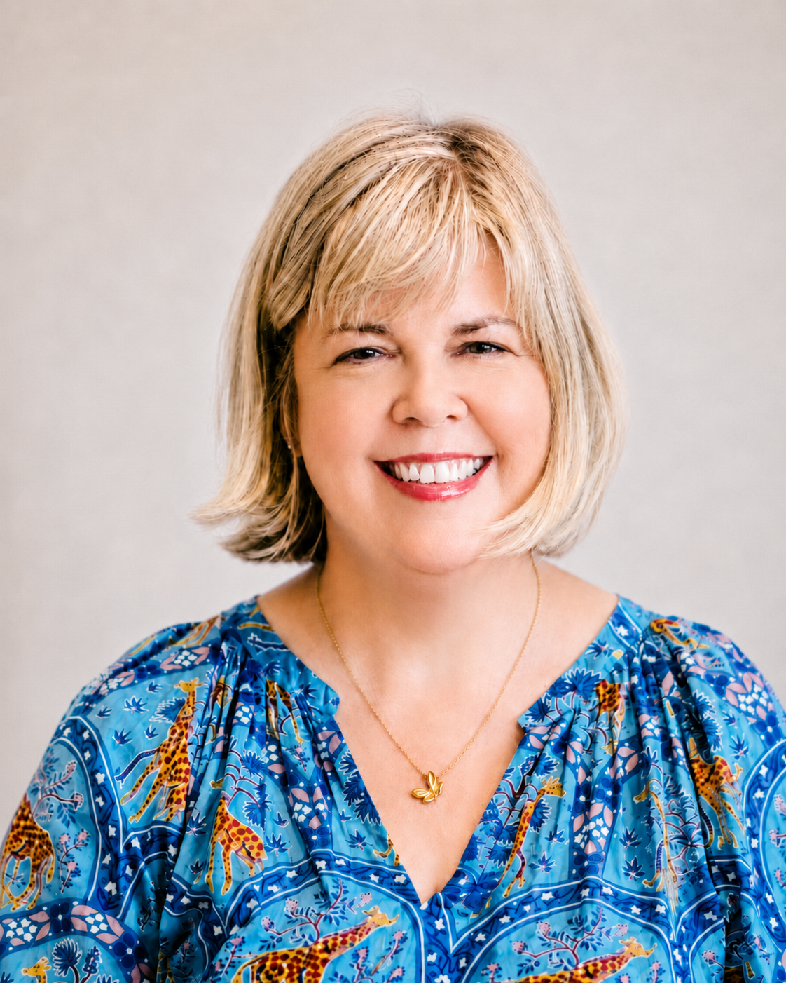 Smiling blonde woman with short hair wearing a blue patterned top and a gold necklace with a leaf pendant against a plain light background.