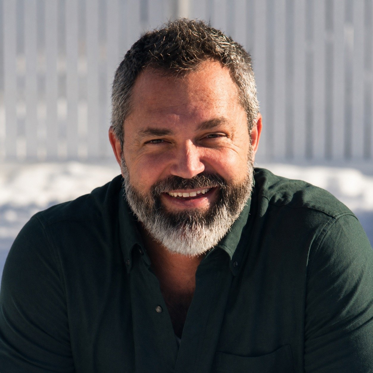 A smiling middle-aged man with a beard and short hair, wearing a dark green shirt, outdoors in snowy weather with a blurred fence in the background.