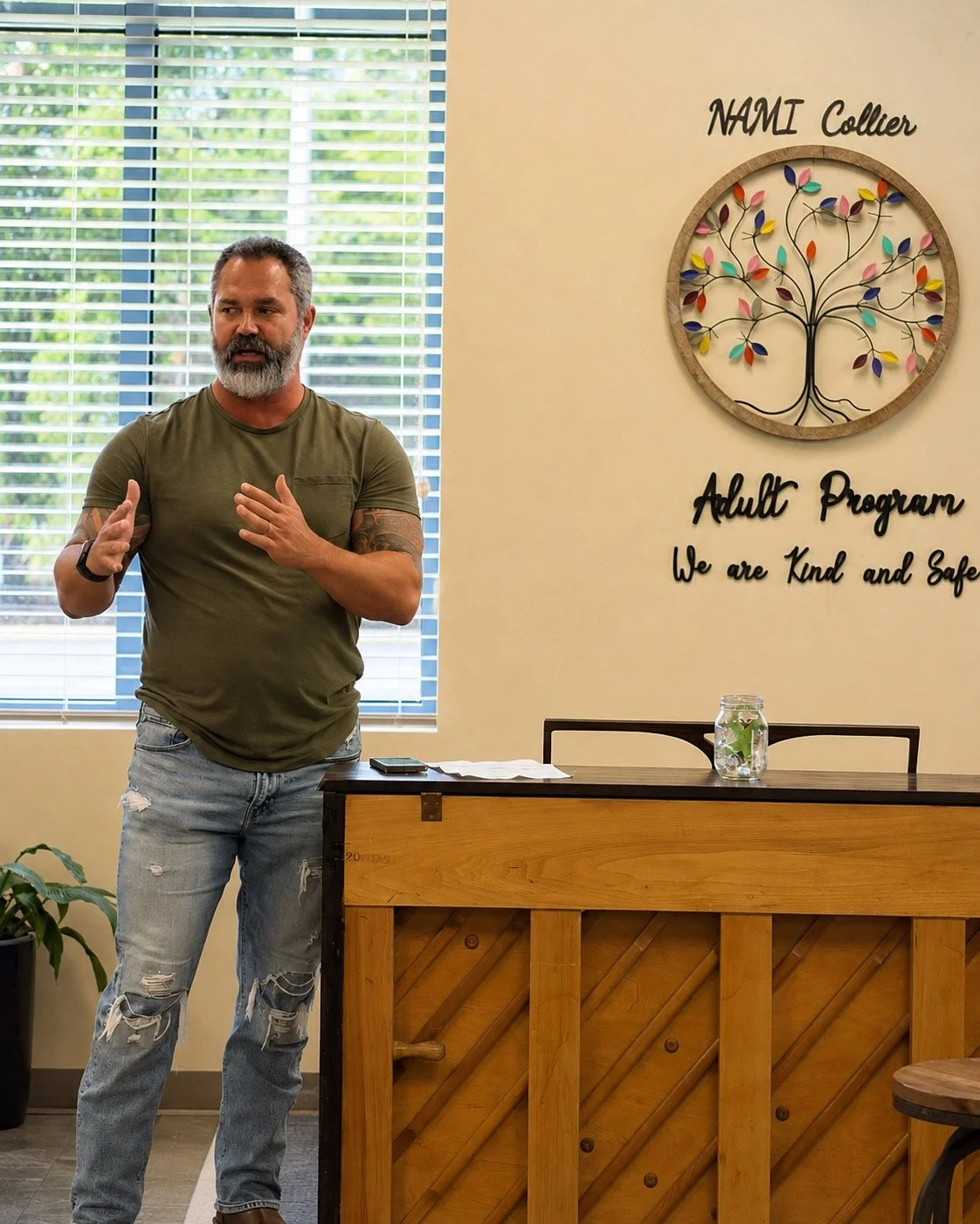 A man with a beard and tattoos on his right arm, wearing a green t-shirt and ripped jeans, stands in front of a window with blinds, speaking or giving a presentation at an adult program called 'NAMI Collier.' Behind him is a wall with a colorful tree wreath and a sign that says 'Adult Program. We are Kind and Safe.' A table with a jar of water and some papers is in front of him.