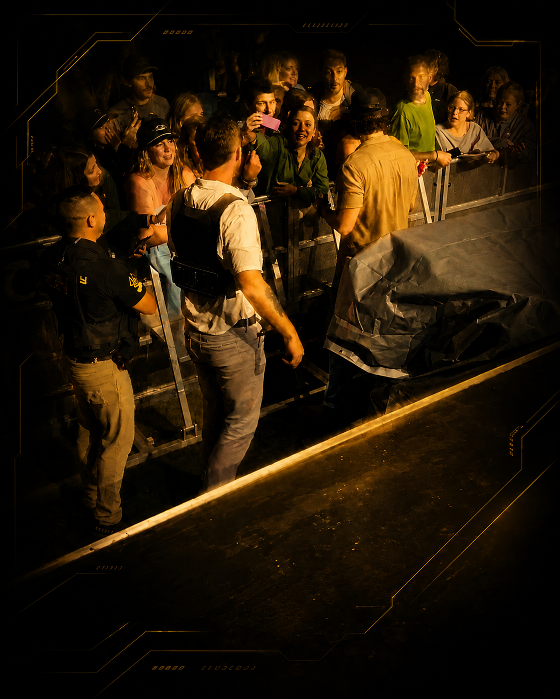 Group of fans gathering behind barricade, taking photos and talking to a man in a yellow shirt on stage, with Monarch Security agents nearby