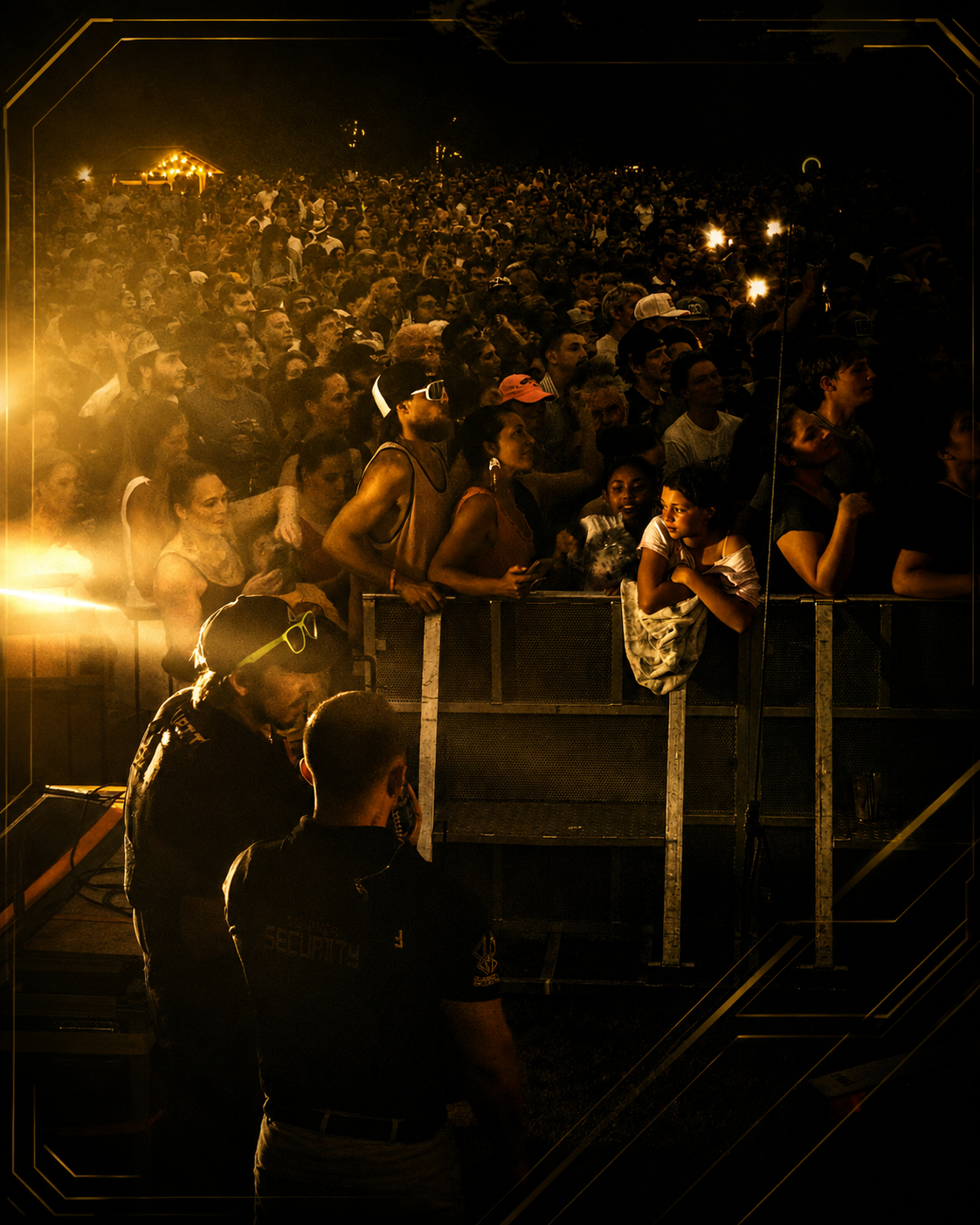 Crowd of people attending a nighttime outdoor event, standing behind a metal barrier, with Monarch security personnel present.