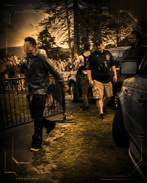 A man being protected by a Monarch Security Agent standing next to a fence with a crowd of people and cars nearby