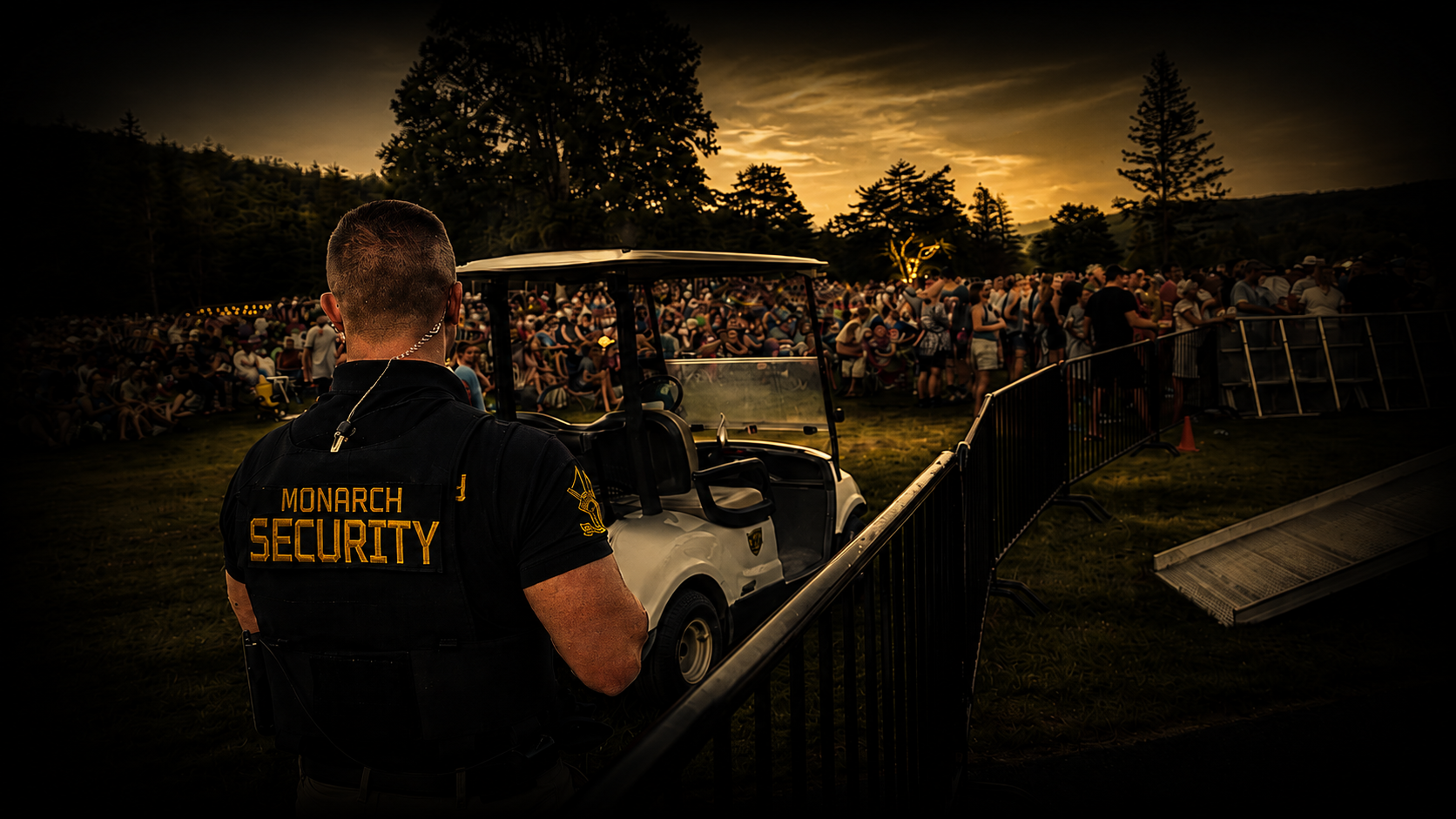 A Monarch security officer wearing a black shirt with yellow 'Monarch Security' text stands in front of a golf cart, overseeing a large crowd of people gathered outdoors