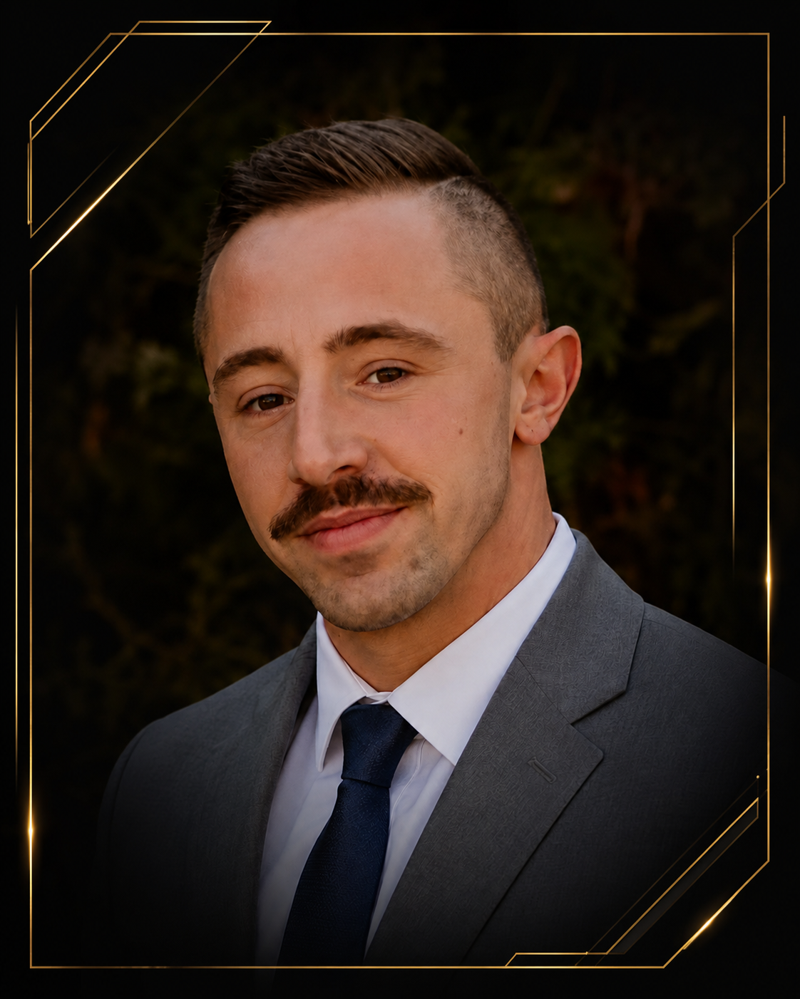 A man with short hair and a mustache wearing a gray suit, white shirt, and navy tie, smiling slightly with a dark background.