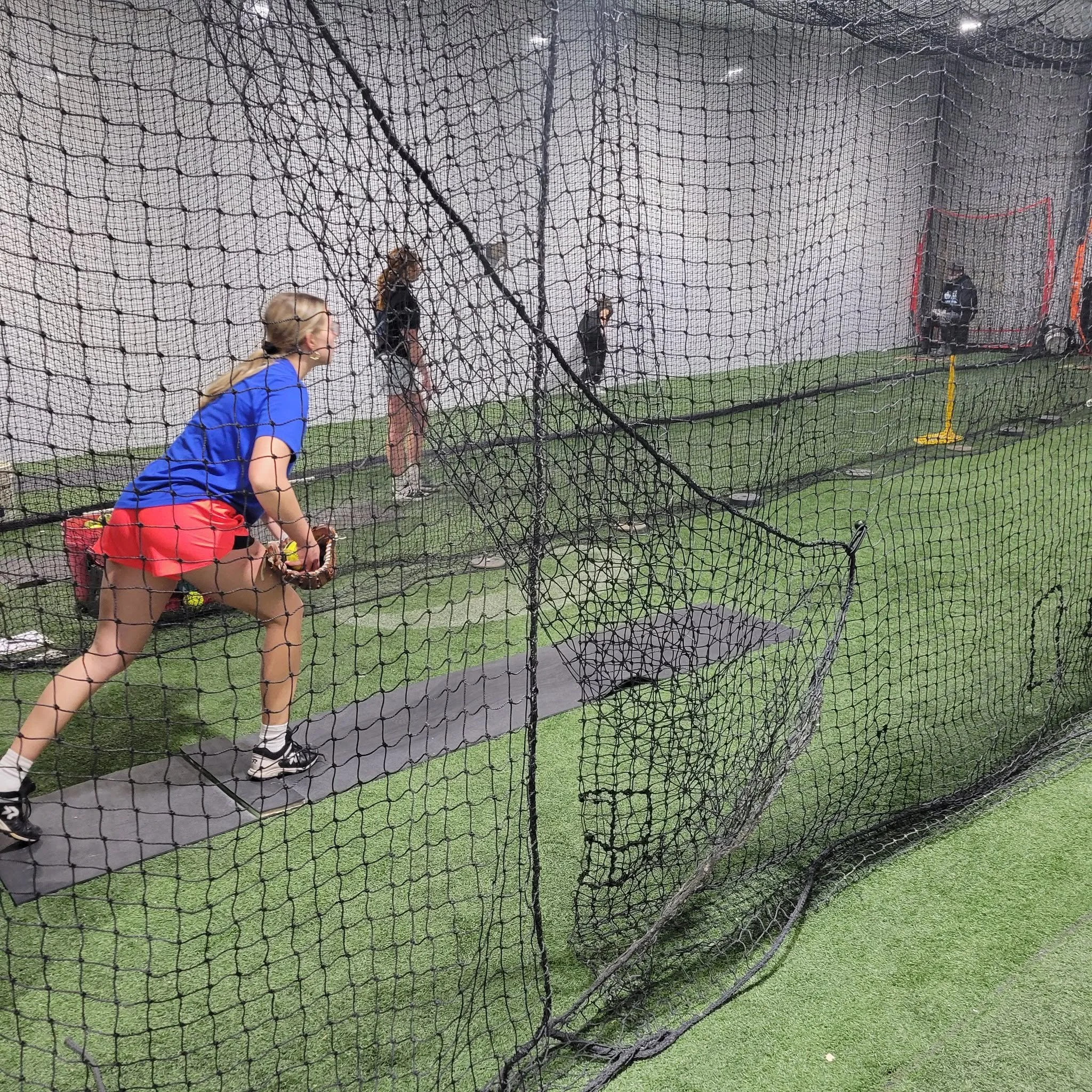 Indoor baseball or softball batting practice area with a net enclosure, three women in athletic clothing practicing, and a coach near a pitching machine in the background.