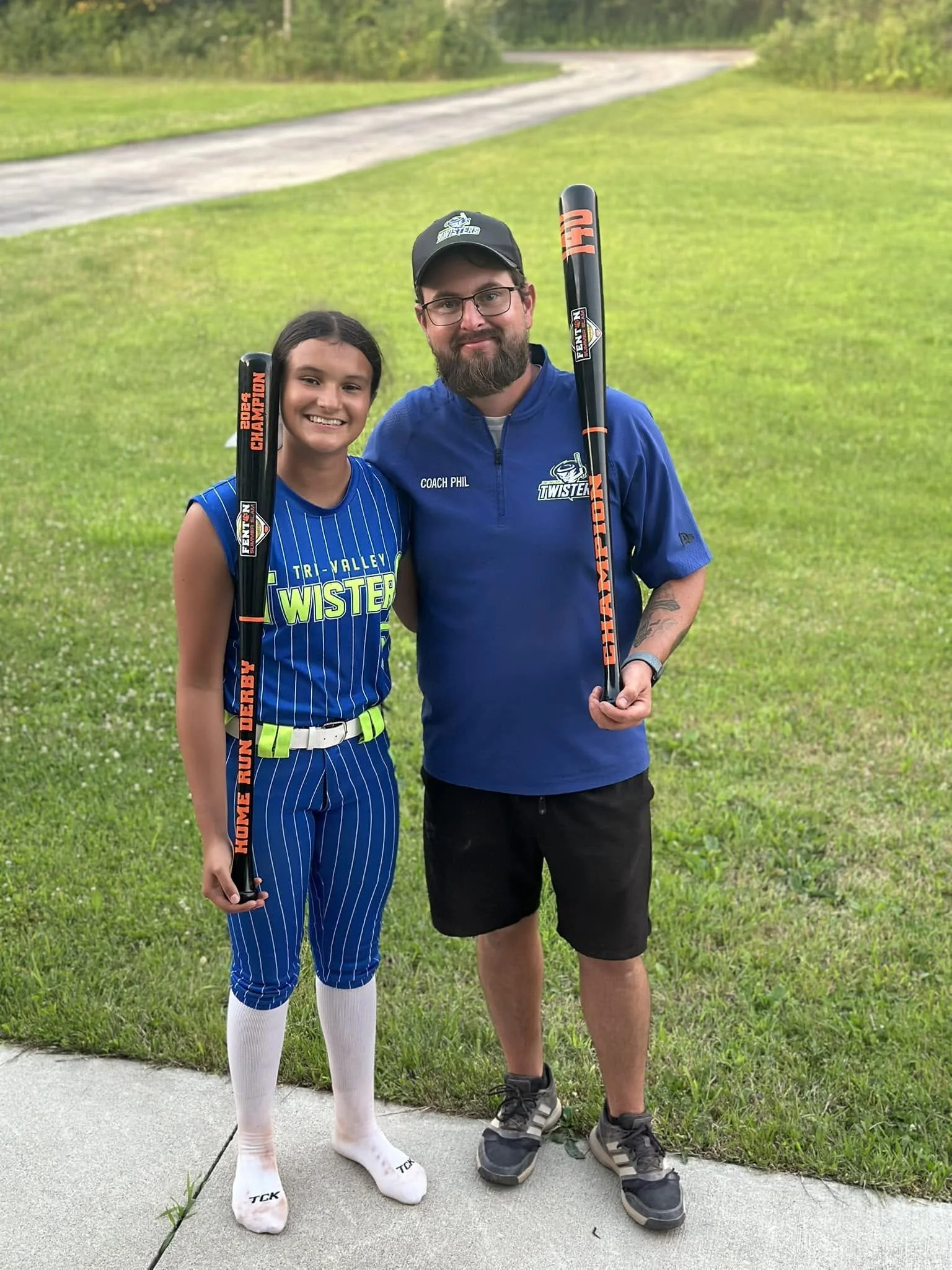 A young girl and an adult man standing outdoors on a sidewalk in a grassy area. The girl is wearing a baseball uniform with a blue jersey that says 'TRI-VALLEY WISTERS,' pinstripe pants, knee-high socks, and holding a baseball bat. The man is dressed in a blue t-shirt with 'COACH PHIL' embroidered on it, black shorts, and gray sneakers, also holding a baseball bat. They are smiling at the camera.