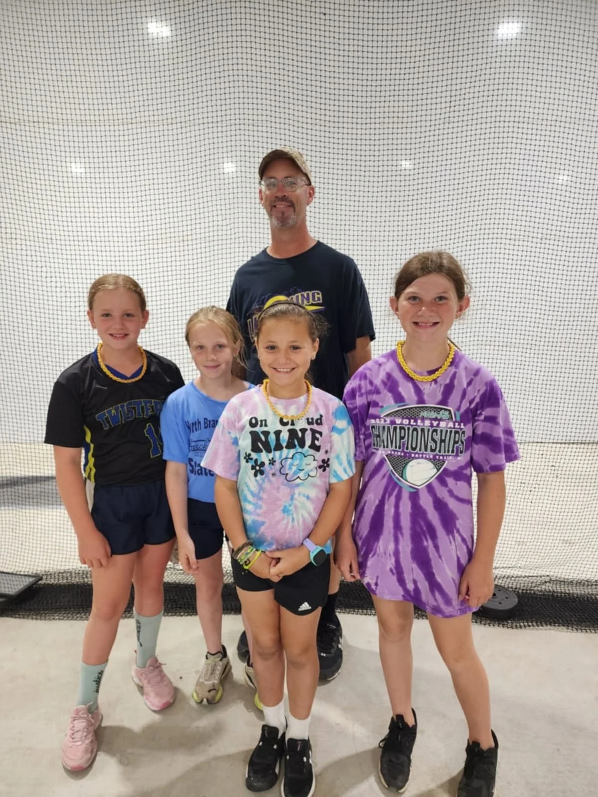 Group of five young girls and one adult man standing inside a gymnasium with a black netted wall behind them. The girls are wearing sports jerseys and casual clothes, and the man is dressed in sportswear. They are smiling and posing for the photo.