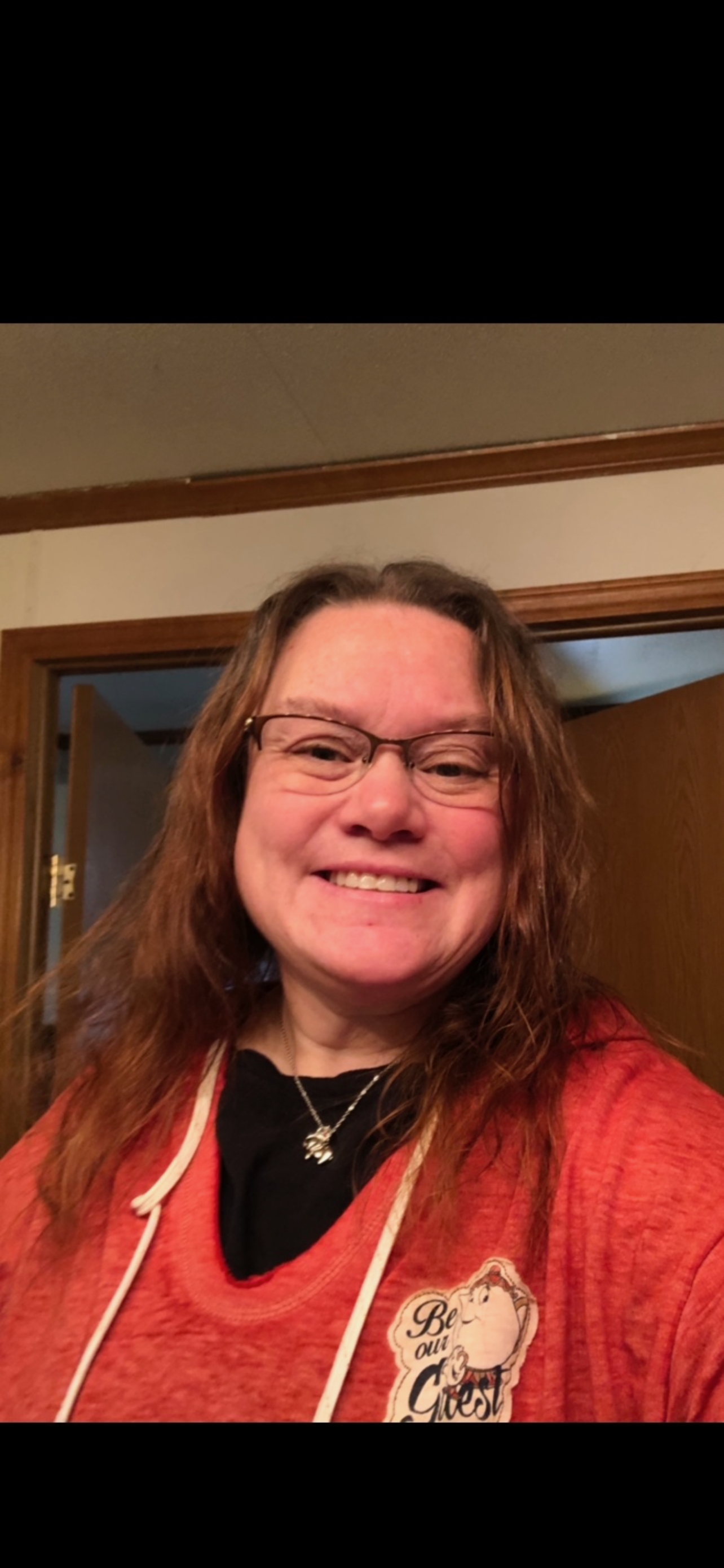 A woman with glasses and long brown hair smiling in a doorway, wearing a red hoodie with a graphic on it.