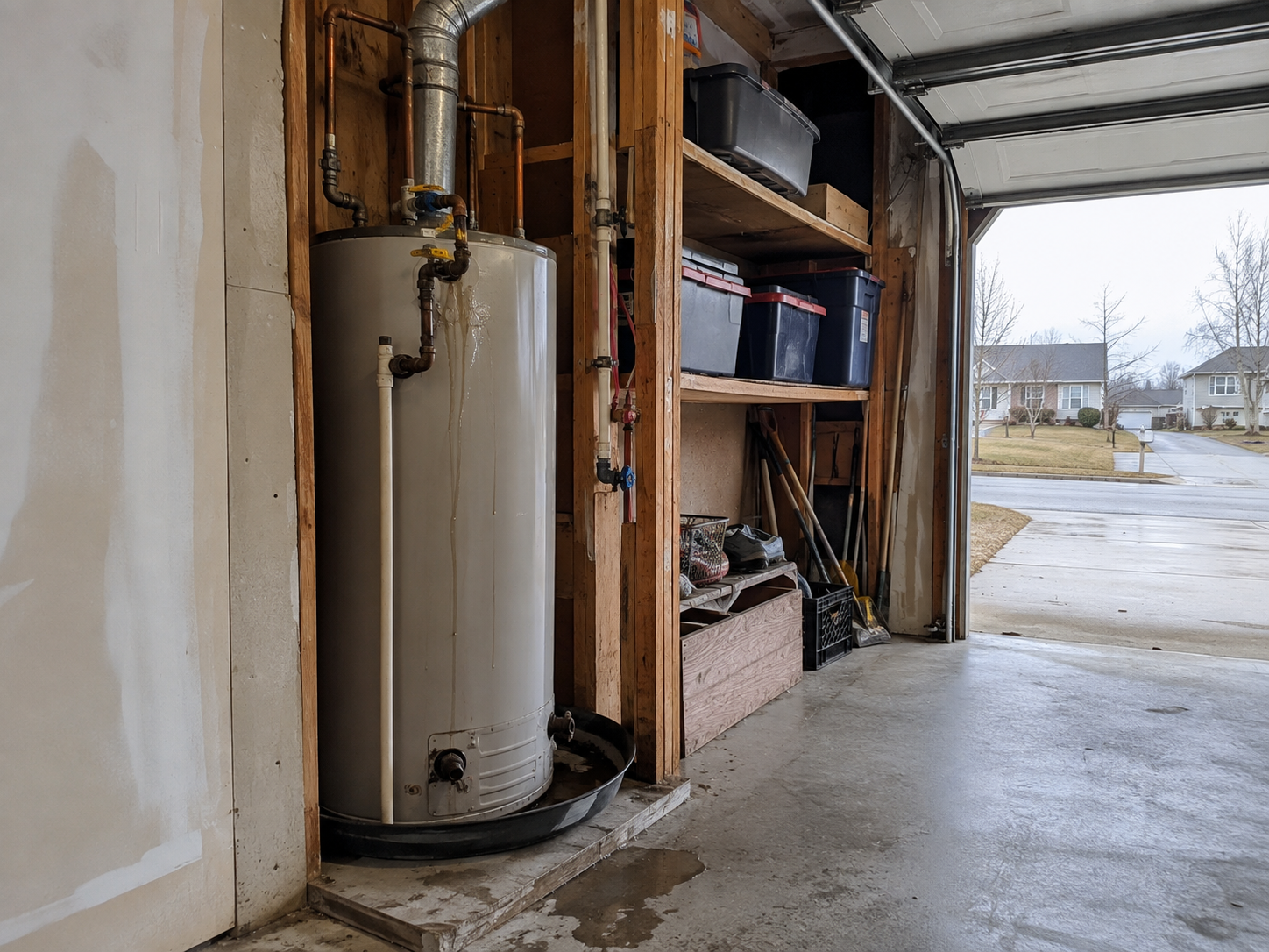 View inside a garage showing a water heater, storage shelves with black storage bins, and outdoor view of neighborhood houses and street.