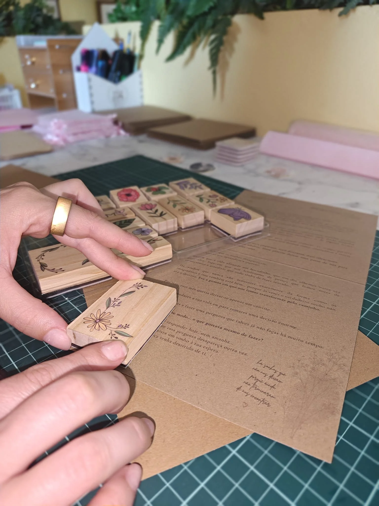 Pessoa a manusear carimbos de madeira com desenhos de flores e palavras, em uma mesa, com arquivos de papel e cartas sobre eles, fundo de parede amarela com plantas penduradas.