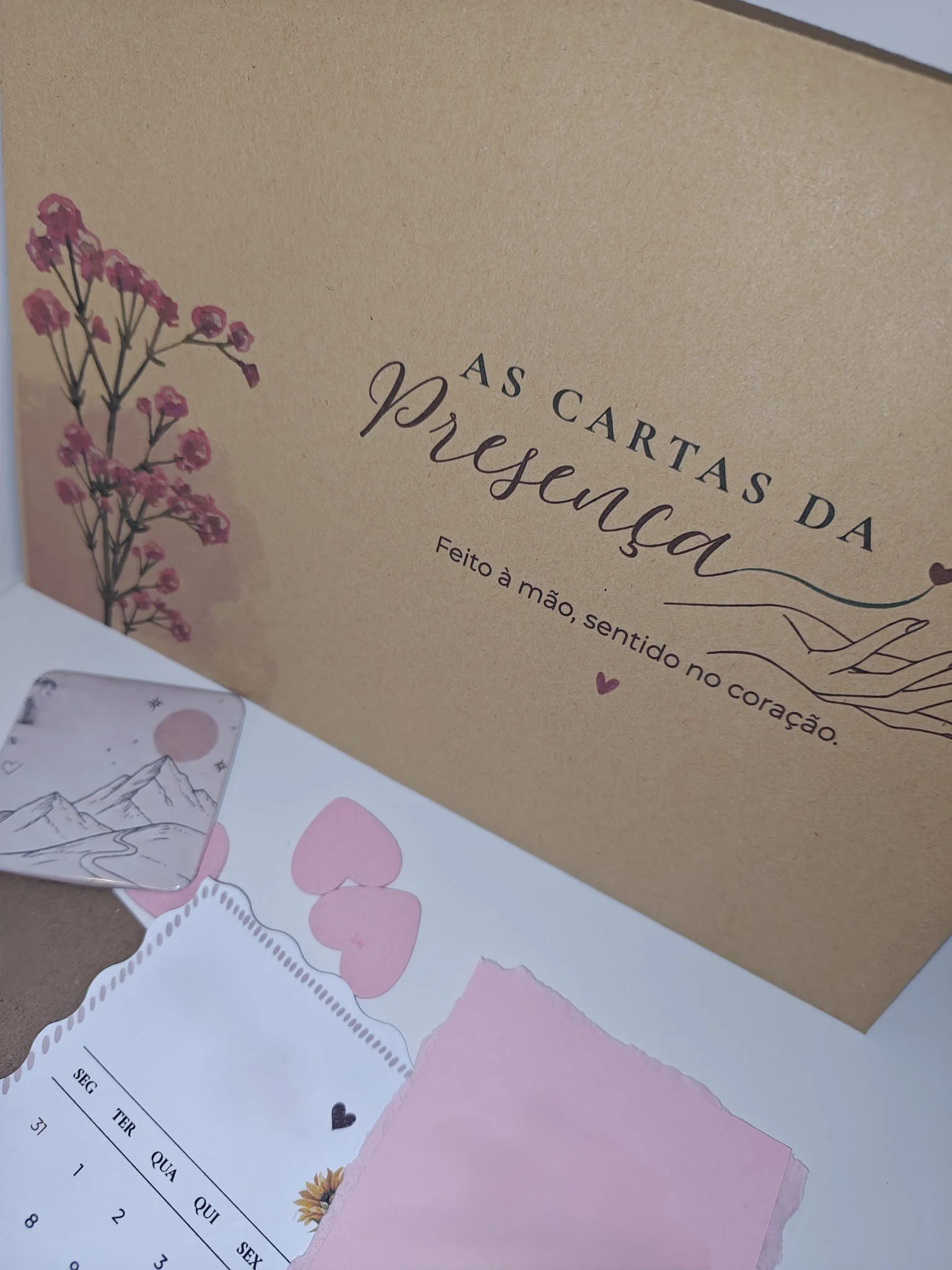 Envelope de papel kraft com desenhos de flores rosas e uma folha de papel rosa por baixo, com parte de uma folha de papel com calendário em português.