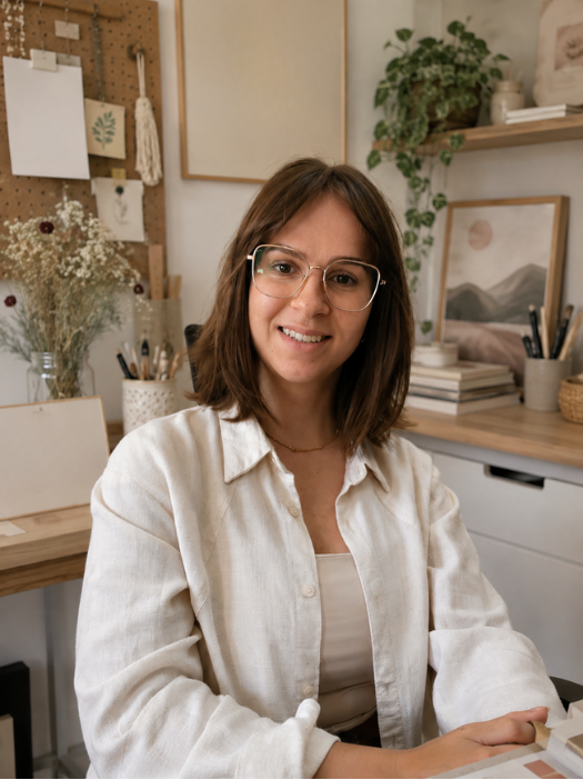 Mulher sorridente com óculos, sentada em um escritório decorado com plantas, quadros e materiais de arte.
