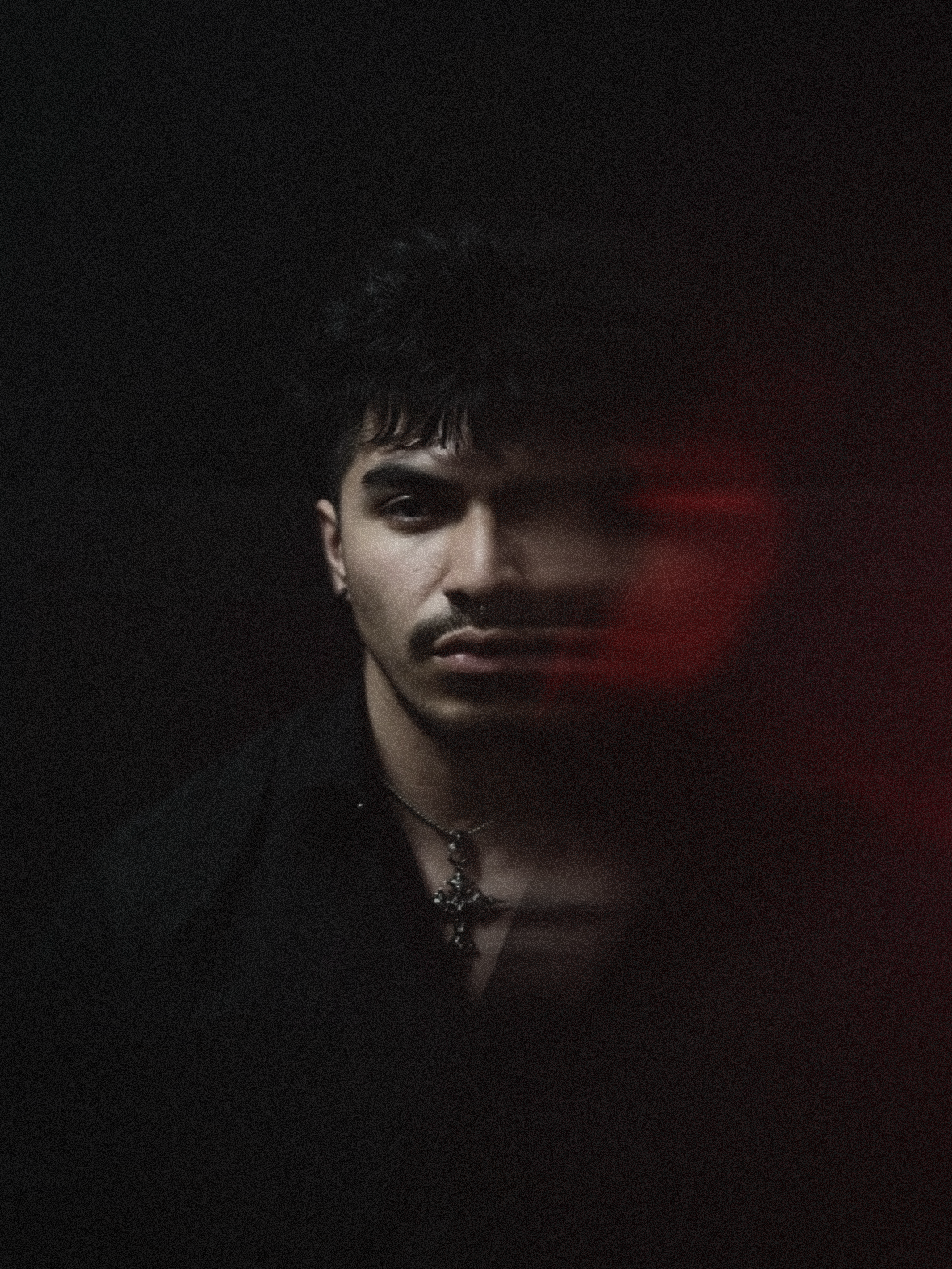 Portrait of a young person with dark hair and makeup, wearing a necklace, in a dark setting with a red light reflection on the right side.
