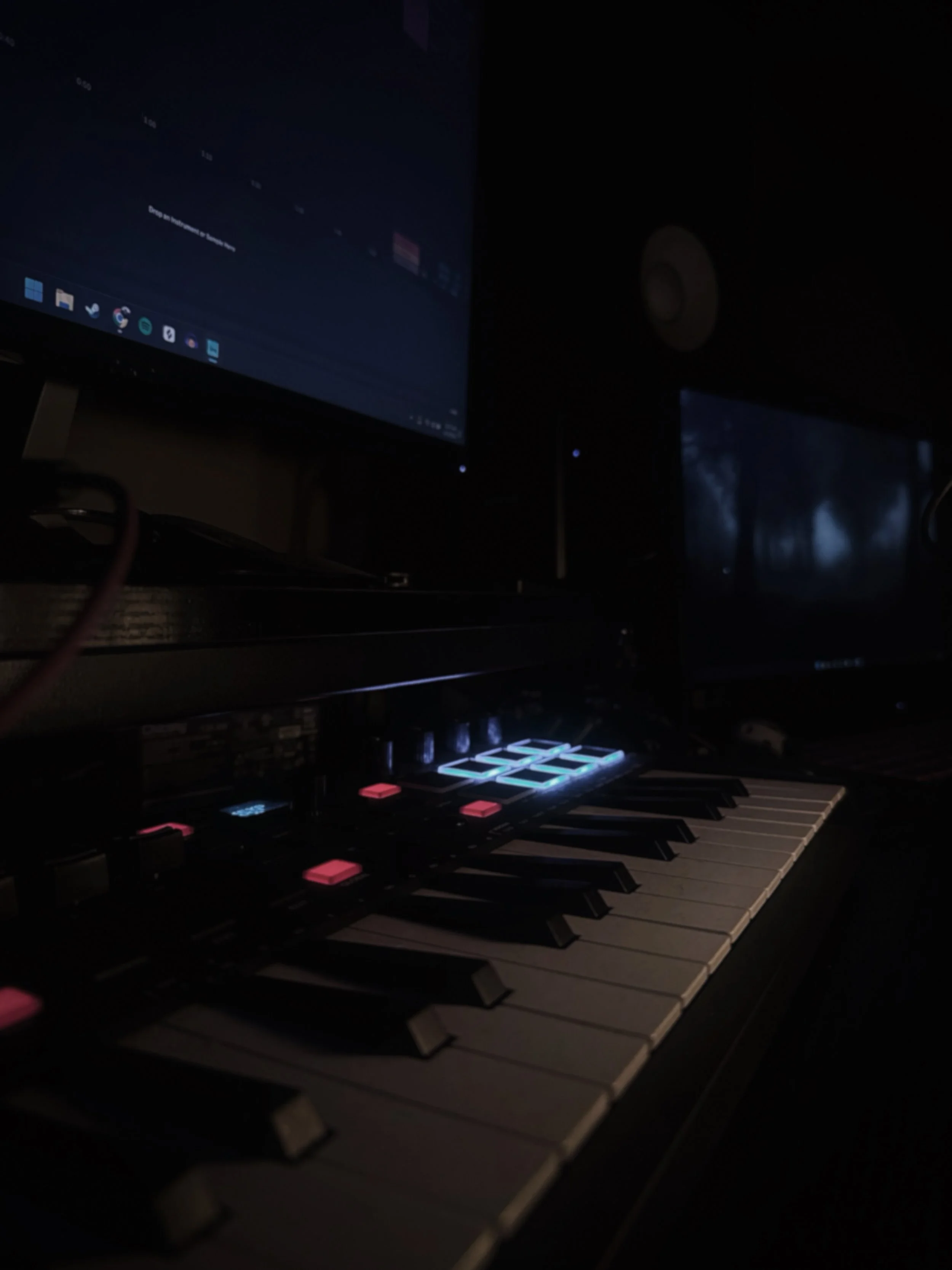 A dark room with a computer monitor, a keyboard with illuminated pink and white keys, and a music production controller with illuminated buttons.