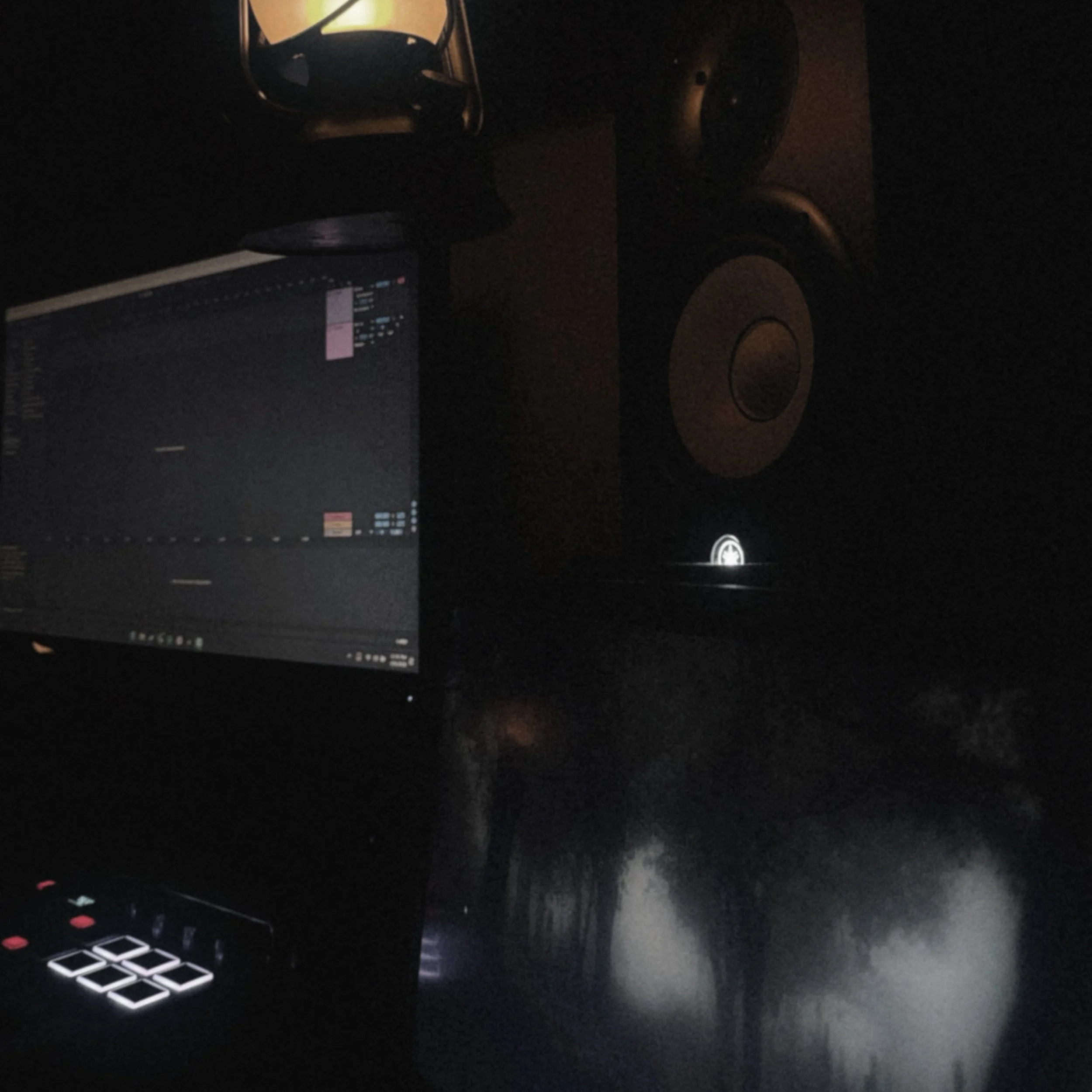 A dimly lit room with a computer screen displaying a digital audio workstation, a makeup mirror with warm lighting, a black chair, some small objects on a desk, a roll of paper towels, and a black desktop speaker.
