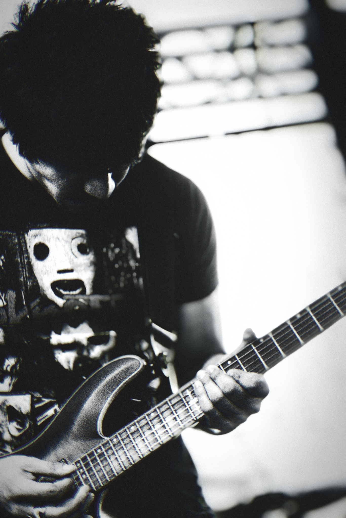 A person with disheveled hair and wearing a skeleton-themed T-shirt is playing an electric guitar. The background shows a window with blinds.
