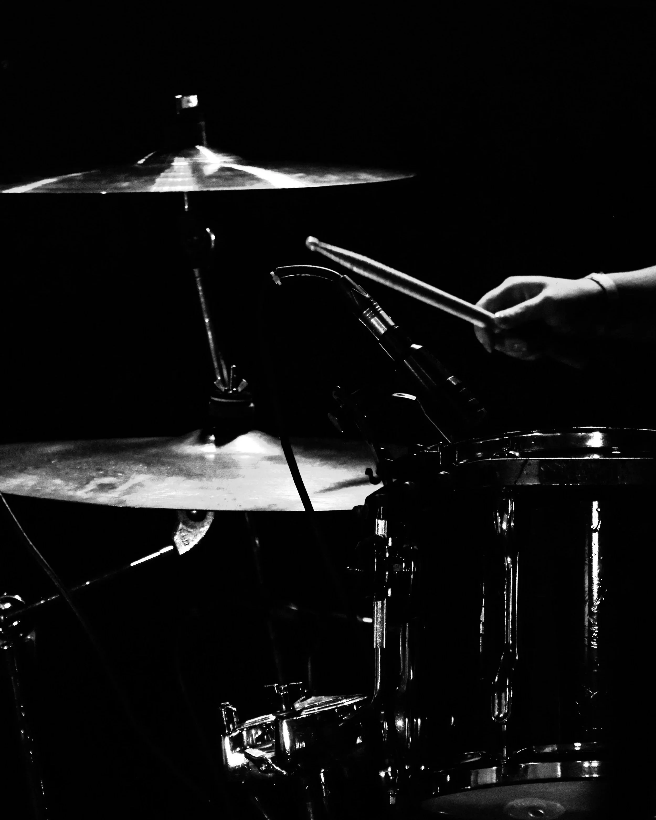 Black and white photo of a drum set being played in a dark setting, showing a cymbal and a hand holding drumsticks.