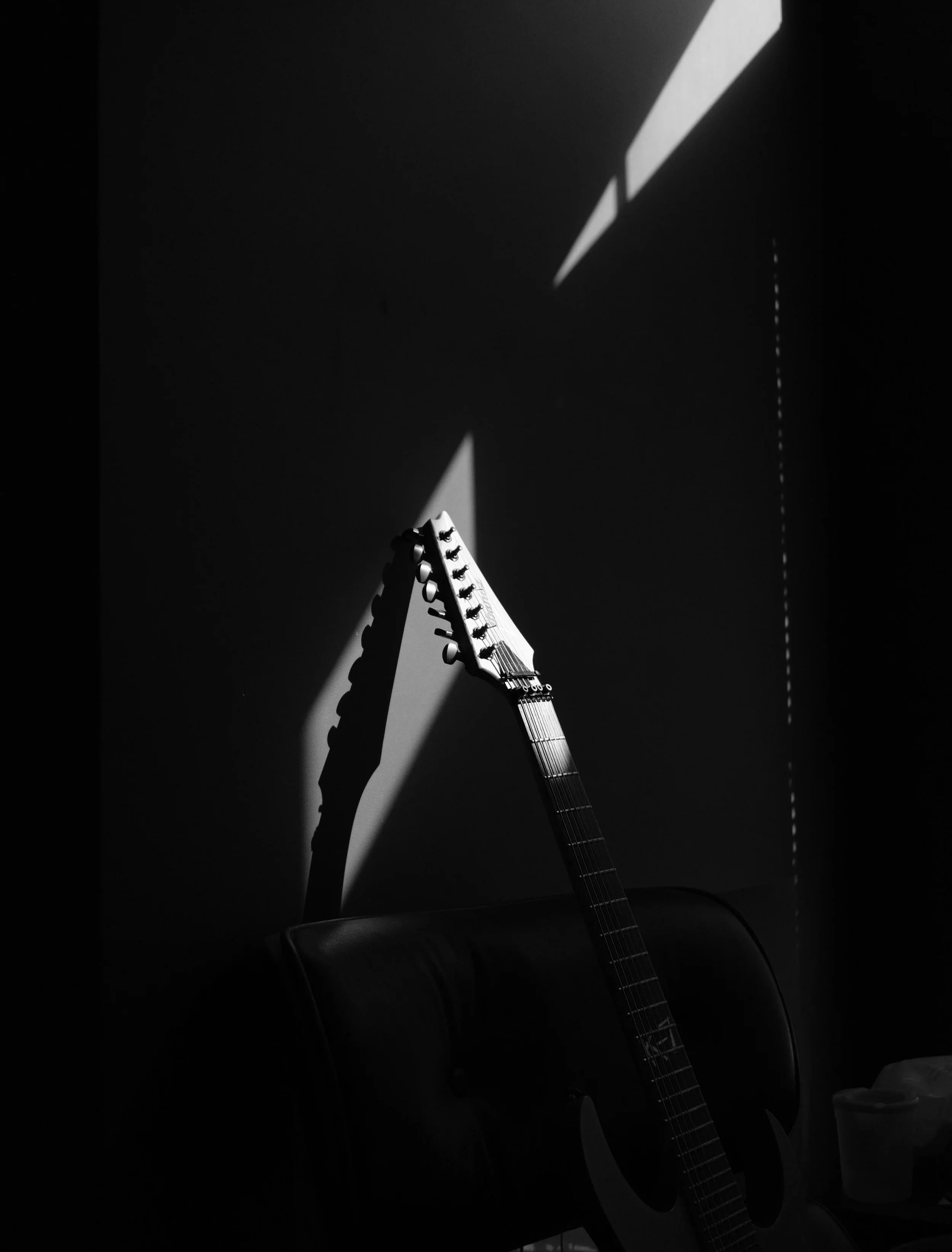 Black and white photo of an electric guitar with a shadow cast on the wall behind it, with a row of chairs and a cup visible at the bottom of the image.