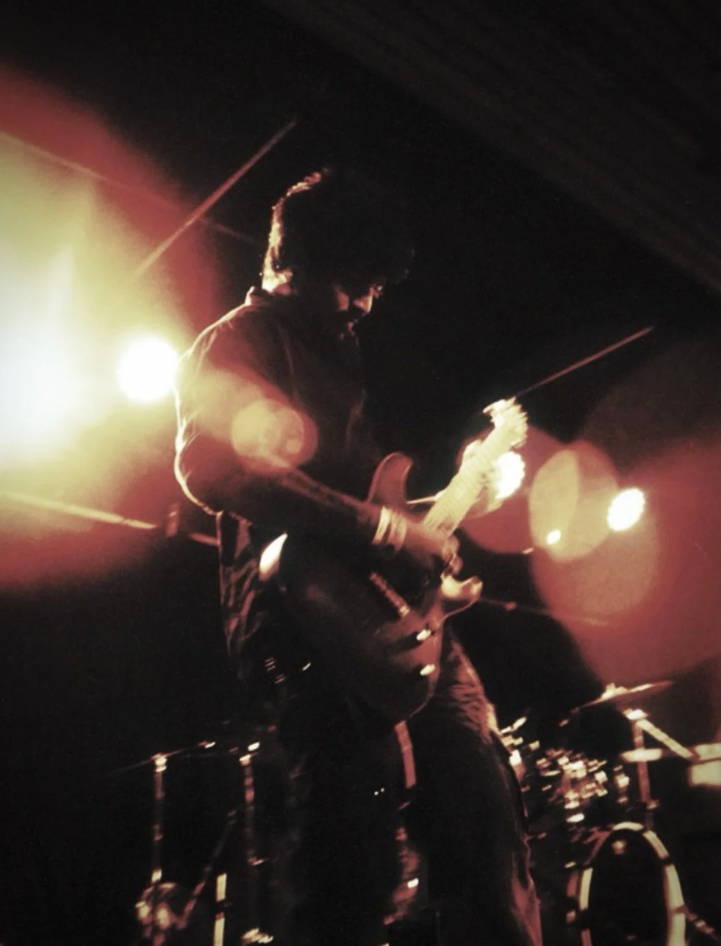 Musician playing electric guitar on stage in dark with bright stage lights behind him.
