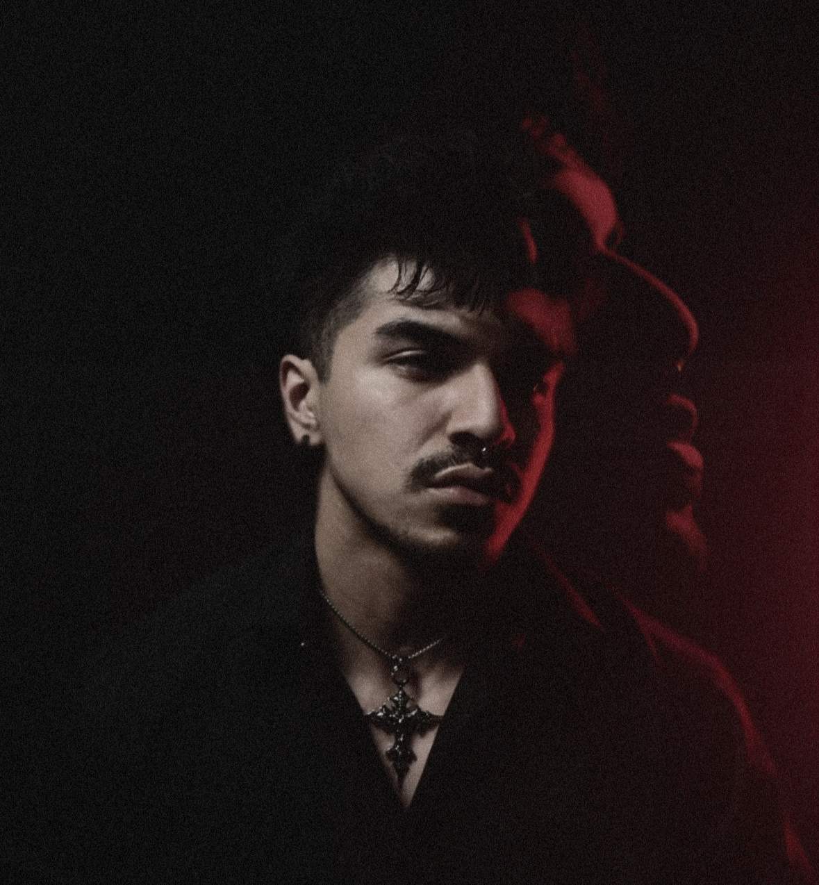 A young man with dark hair and a mustache, wearing jewelry, is illuminated by red and soft light against a dark background, with a faint overlay of his profile in the backdrop.
