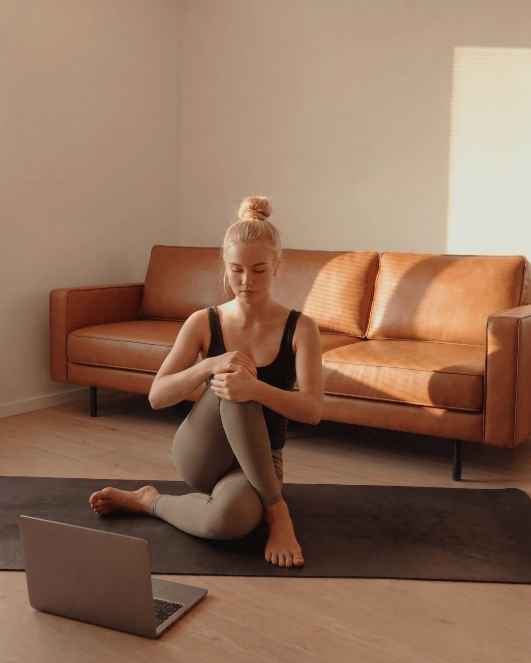A woman practicing yoga on a mat in a room with a tan sofa and wooden floor, sunlight streaming through blinds, looking at her knee.