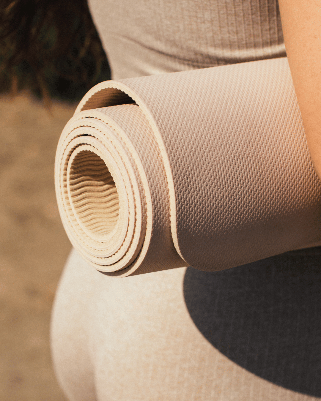 Person holding a rolled-up beige yoga mat outdoors.