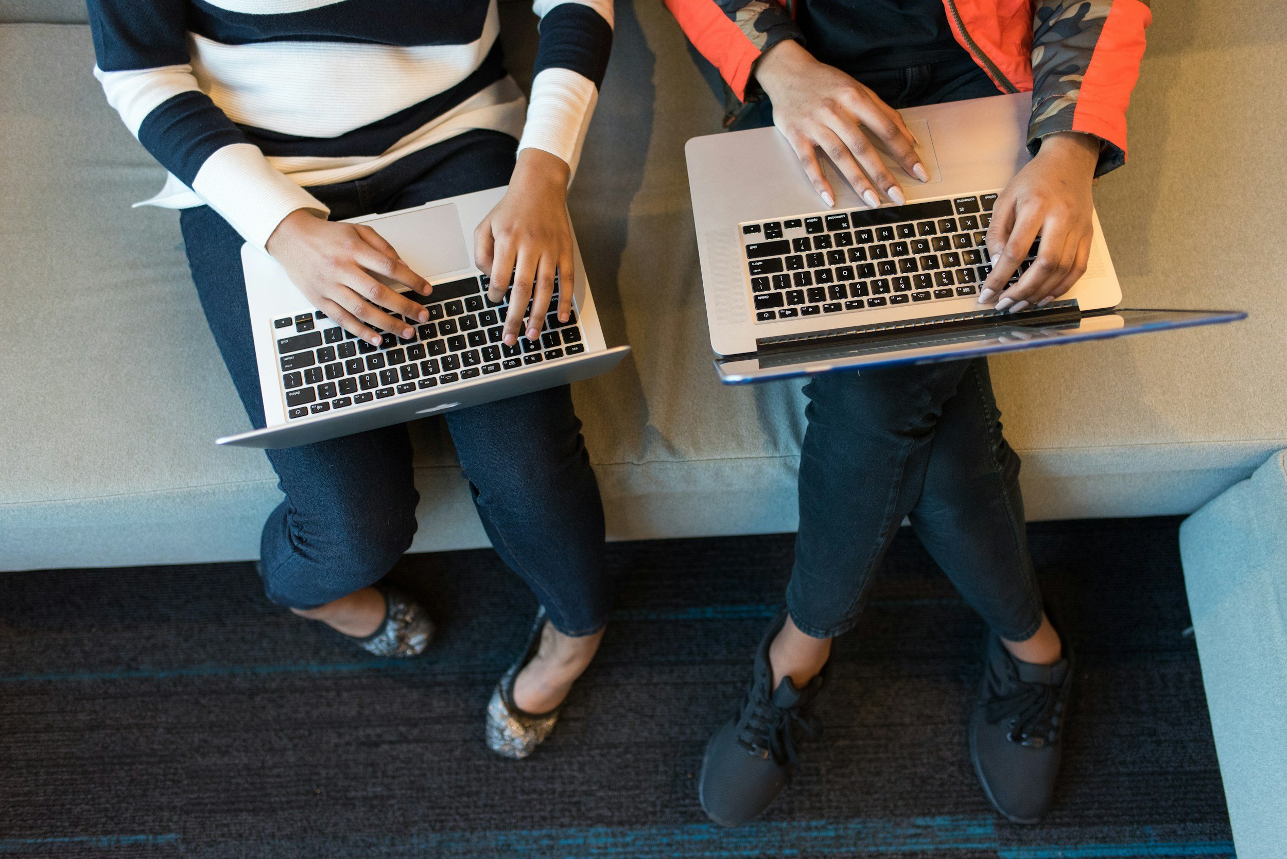 Two people sitting close together on a bench, using laptops. One is wearing a white and navy striped long-sleeve shirt with jeans and leopard-print flats, while the other is wearing a black shirt, an orange and camo jacket, black jeans, and black sneakers.