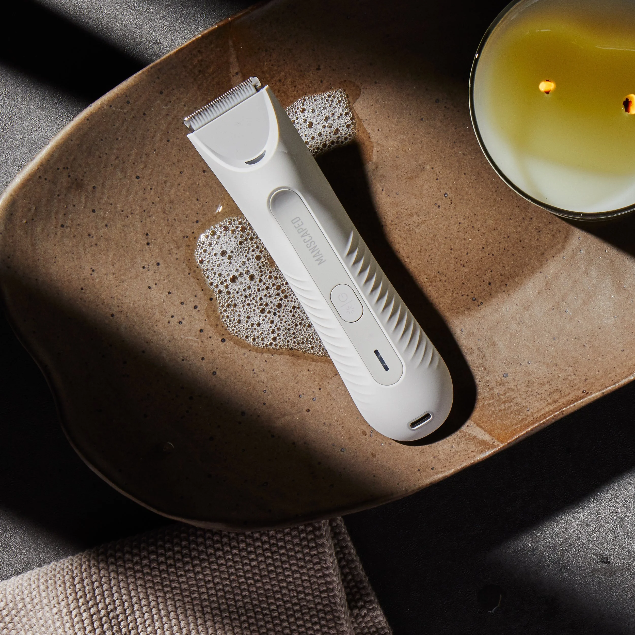 A white electric hair trimmer lying on a brown textured surface with a bowl of liquid and a beige and brown knitted hat nearby.