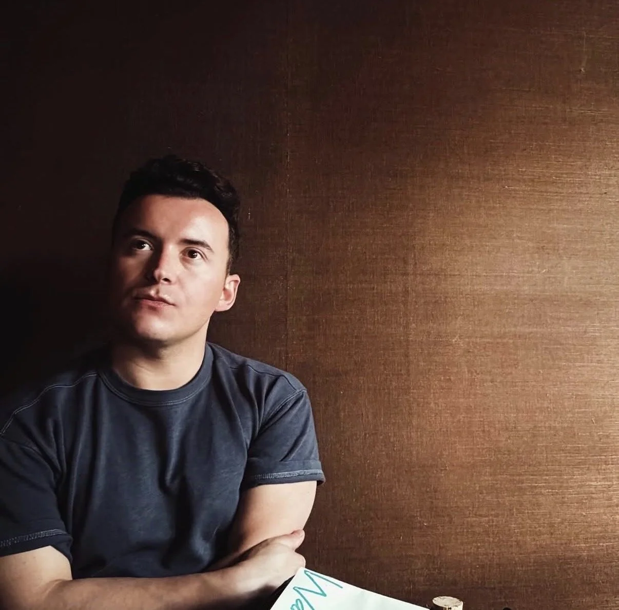 A young man with dark hair sitting at a table against a brown textured wall, wearing a dark gray t-shirt, with his arms crossed on the table.