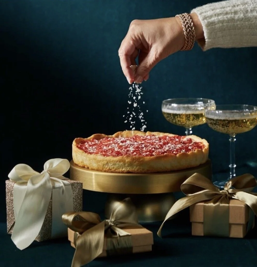 Person sprinkling sugar on a strawberry cheesecake, with two glasses of champagne and three wrapped gifts surrounding it.