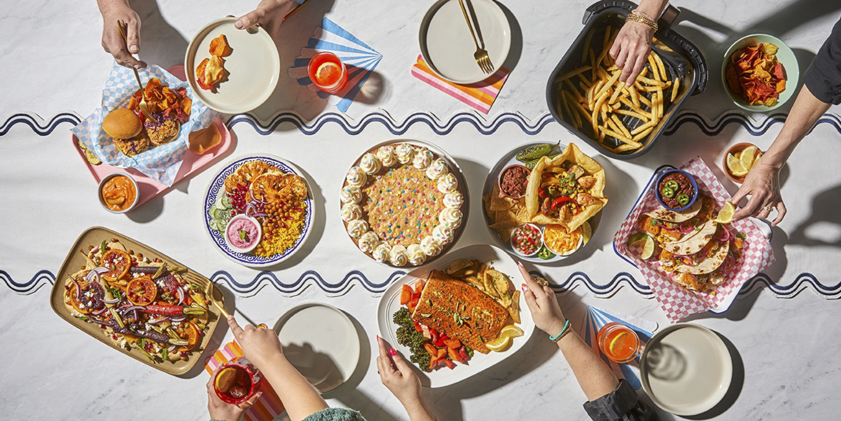 Overhead view of a festive meal with various dishes, including tacos, fried chicken, fish, salad, chili, and desserts, with people reaching for food and drinks on a white table.