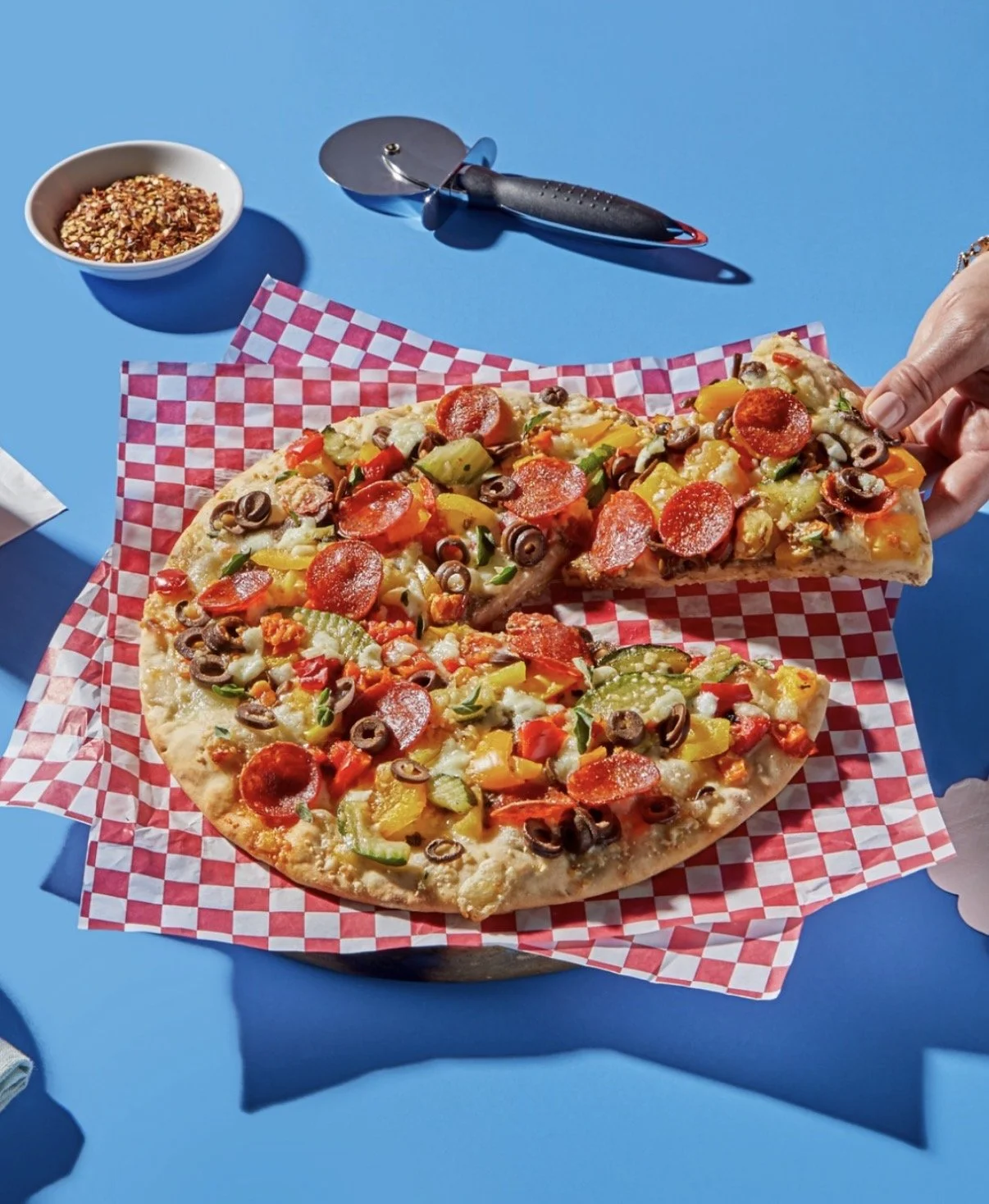 A pizza with various toppings including pepperoni, black olives, green peppers, and yellow peppers on a red checkered paper. A person is holding a slice, and there is a pizza cutter, a bowl of crushed red pepper, and a bowl of crushed red pepper flak