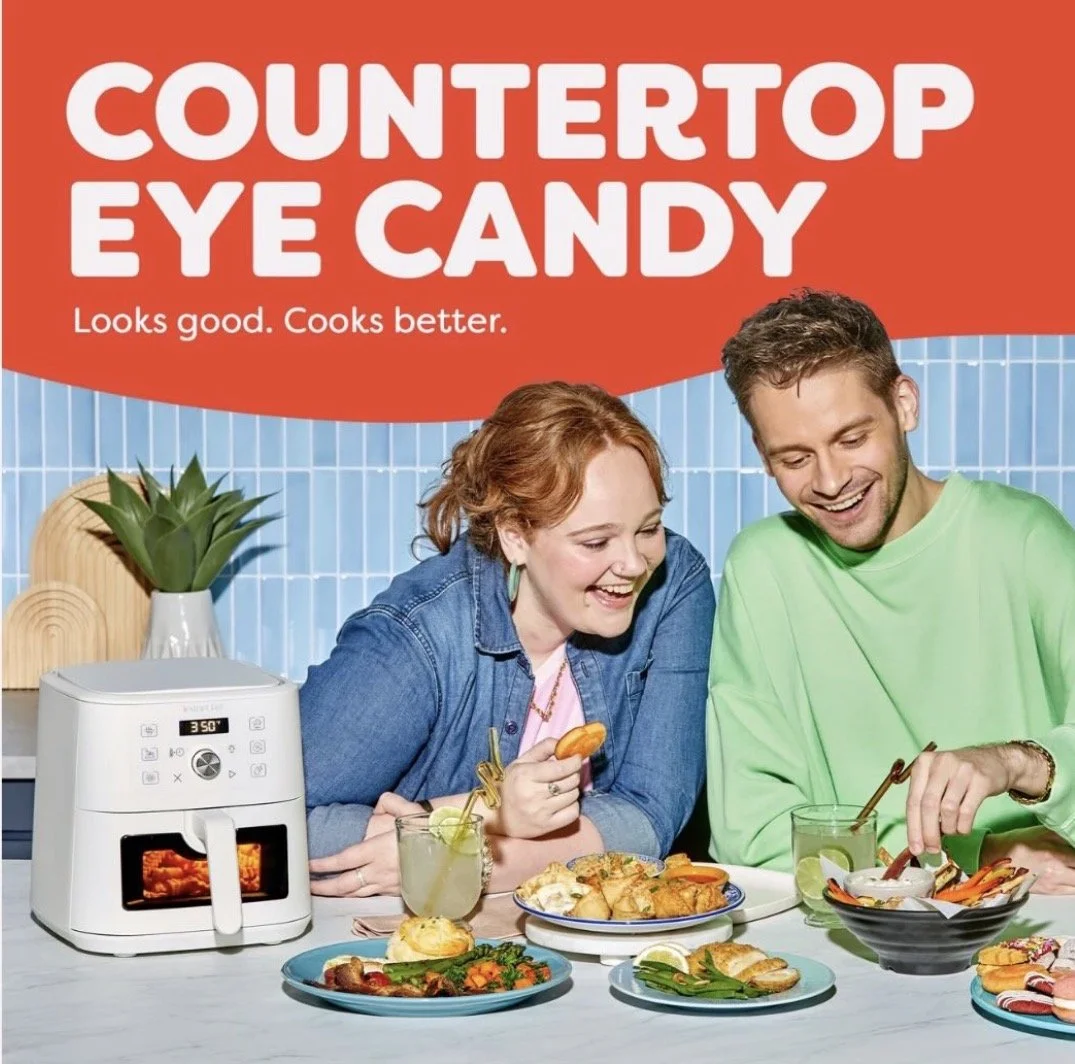 A promotional image for countertop eye candy, showing a happy woman and man cooking and eating together at a kitchen counter with various food dishes and an air fryer in the foreground.