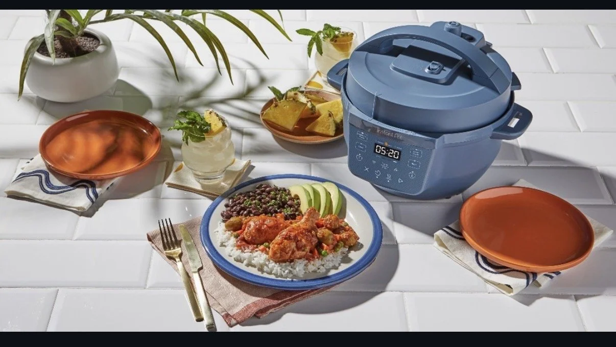 A table set with a plate of rice, beans, and chicken, two orange plates on napkins, a glass of lemonade with mint, a bowl of pineapple chunks, a bowl of potato chips, and a rice cooker in the background.